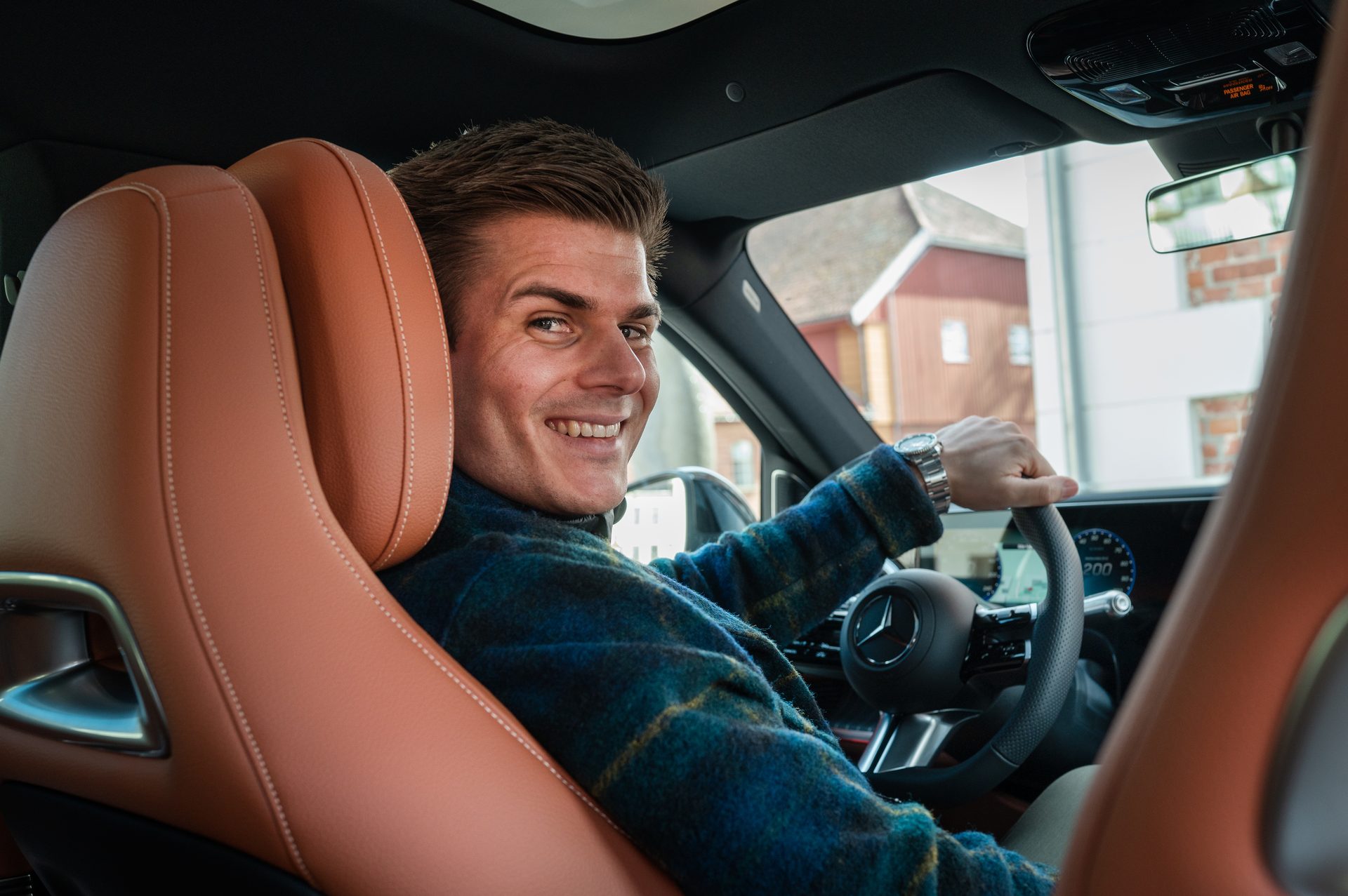 A smiling man in a plaid shirt sits in the driver's seat of a car, looking back.