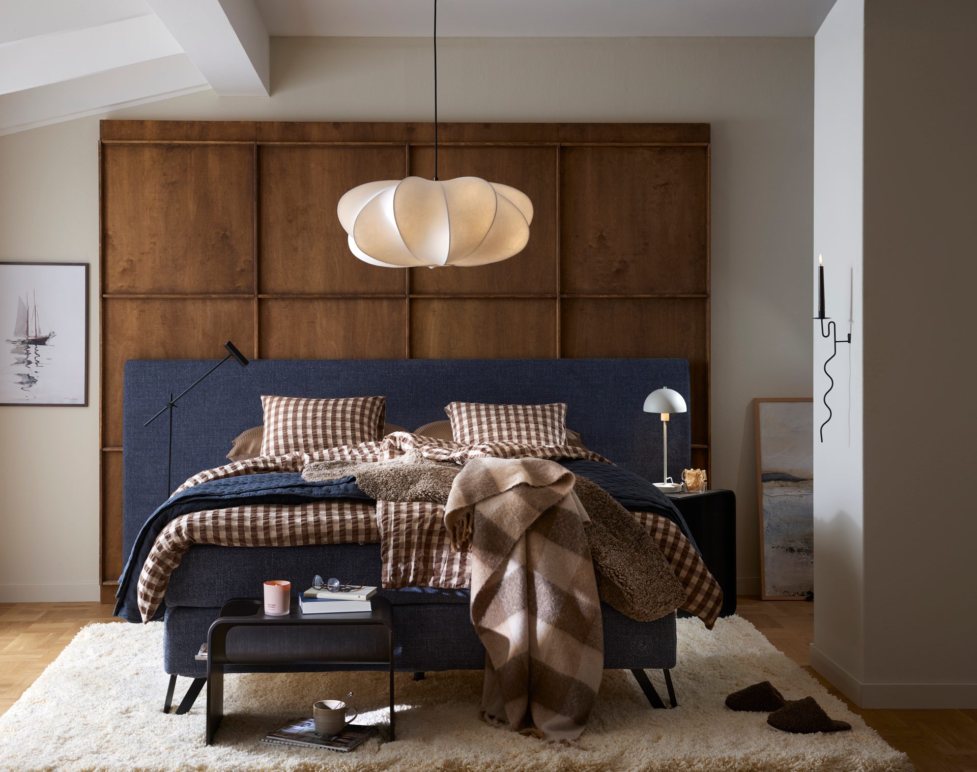 A cozy bedroom with a blue bed, brown checkered bedding, a large white pendant light, and wooden wall panels.