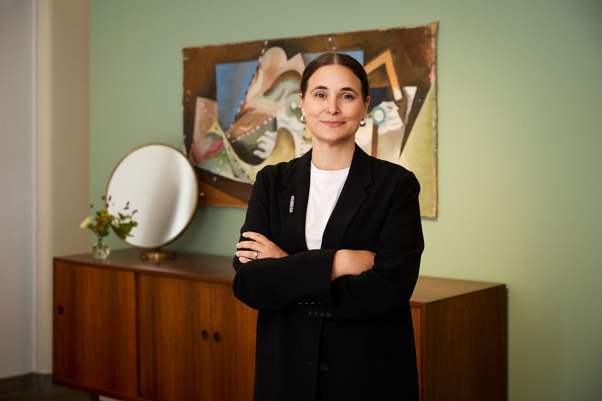 Smiling woman in black blazer, arms crossed, against an abstract painting and wooden sideboard.