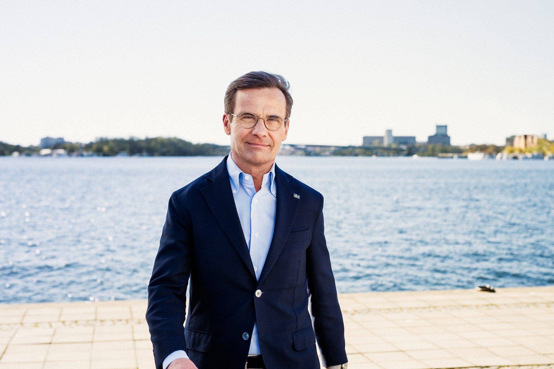 A smiling man in a suit and glasses stands by a body of water with a city skyline in the background.