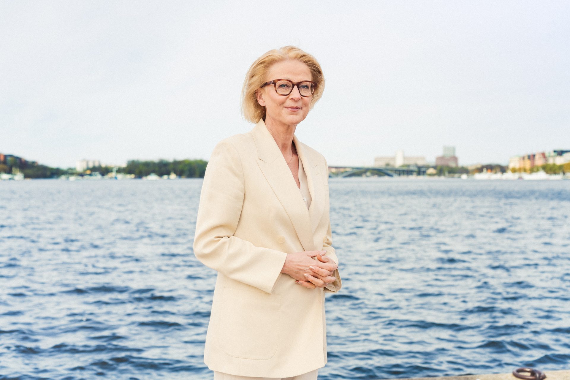A woman in a cream suit and glasses stands by water, with a city skyline in the background.
