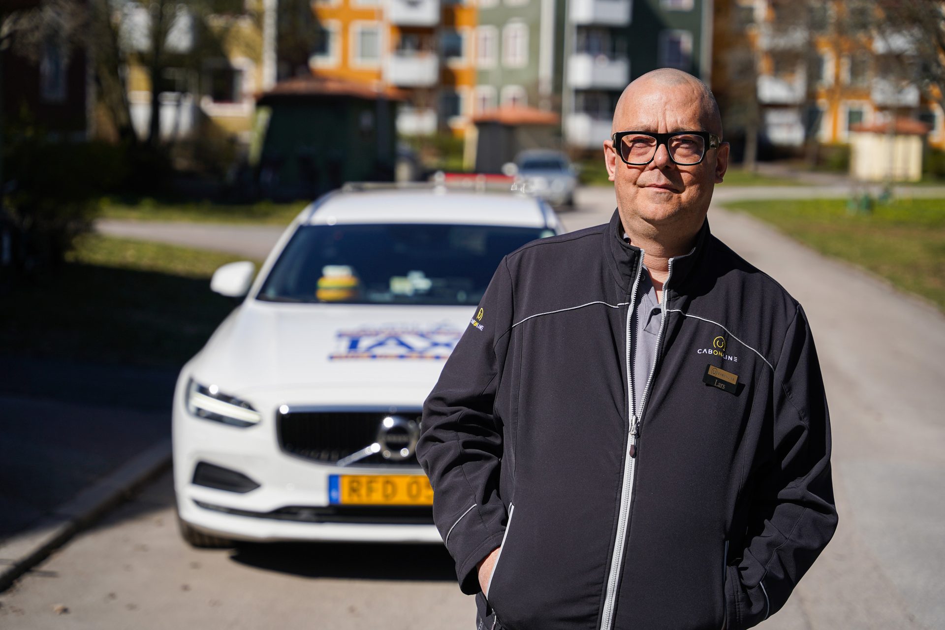 A bald man in glasses and a black jacket stands proudly beside his white taxi car on a sunny day.