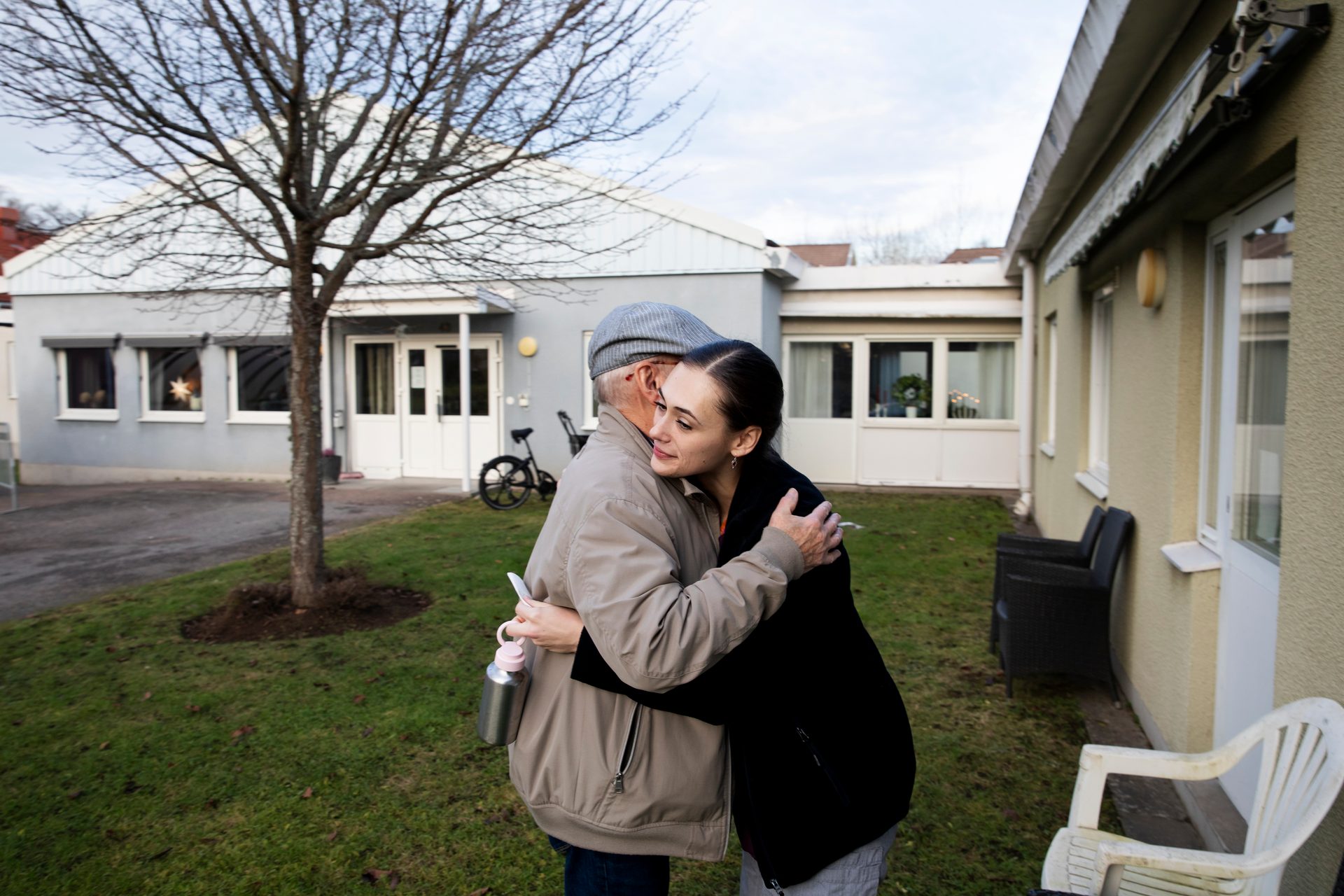 An older man and a young woman hug outdoors, buildings and a tree in the background.