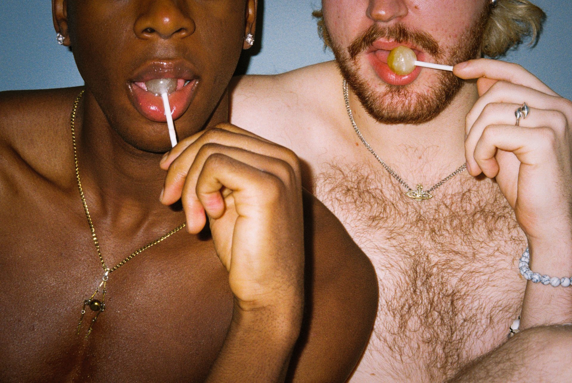 Two shirtless people, one dark-skinned and one light-skinned, with lollipops in their mouths.