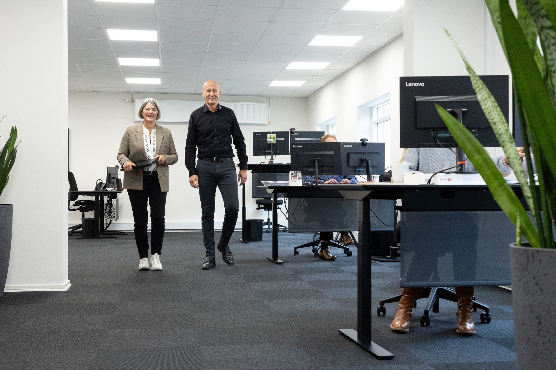 Two smiling colleagues walk through a modern office with people working at desks.