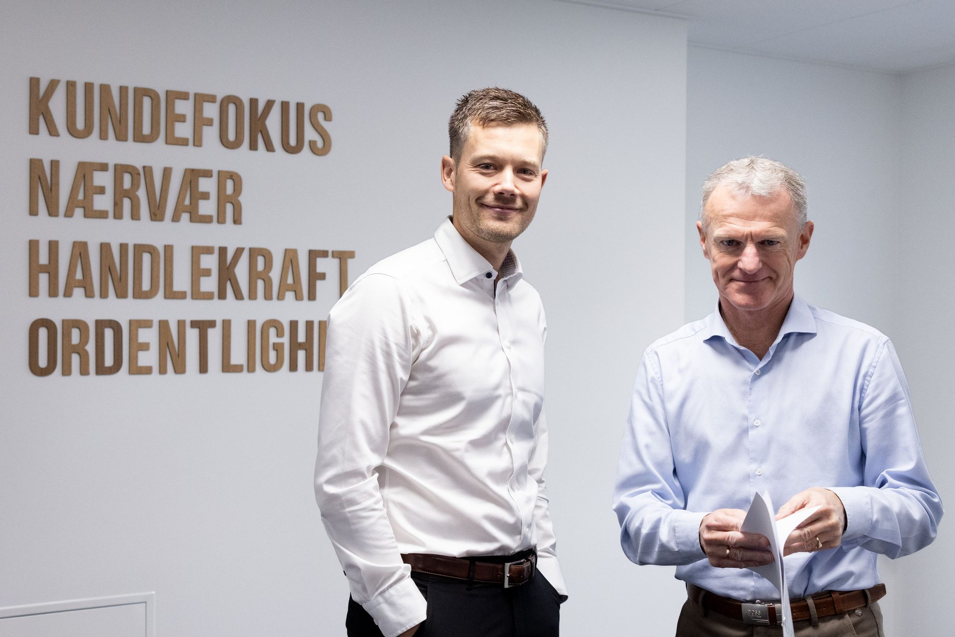 Two men, one smiling, in an office with large Danish words on the wall.