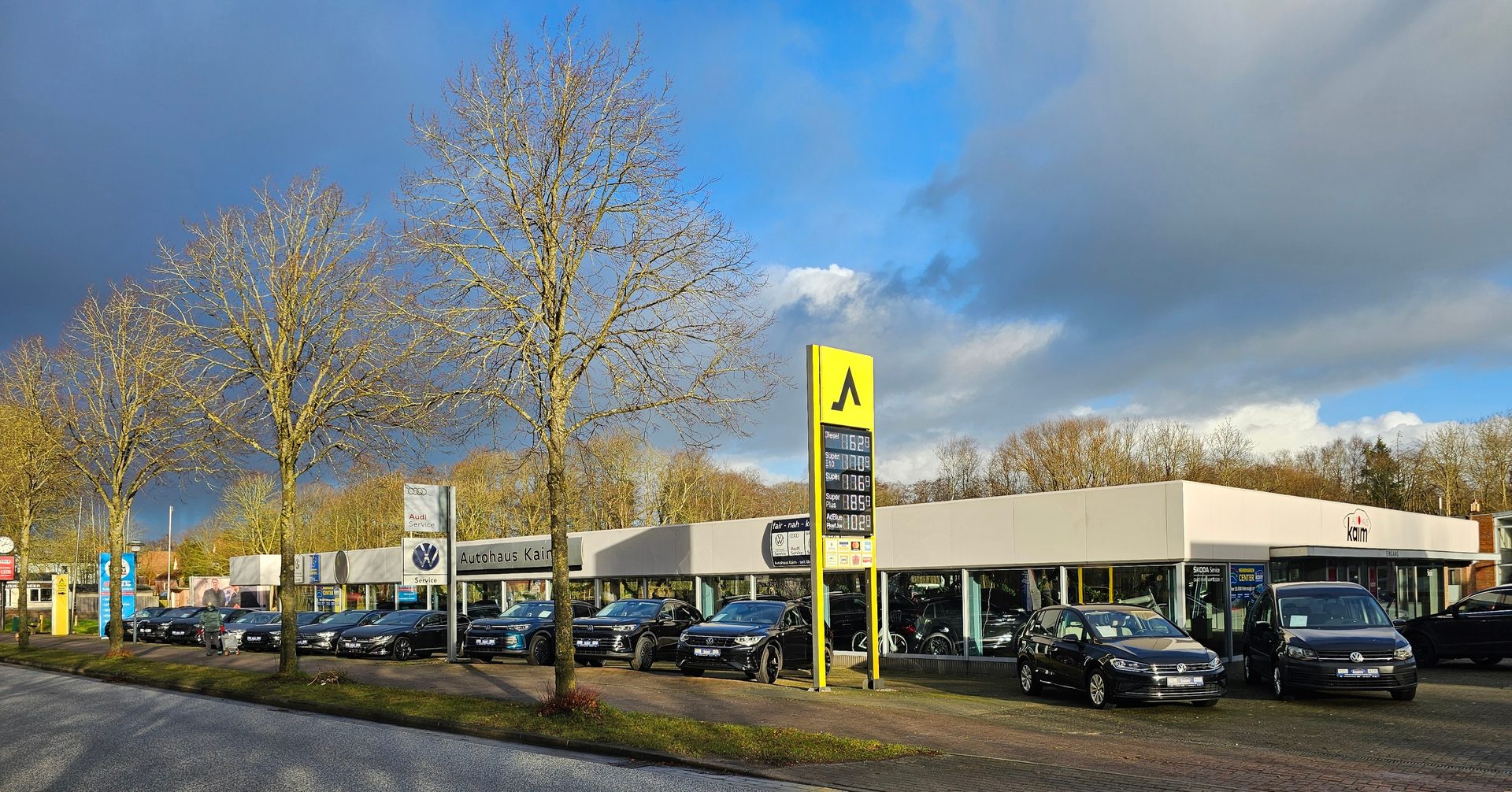 Car dealership (Autohaus Kaim) with cars on display, a gas price sign, and bare trees under a cloudy sky.