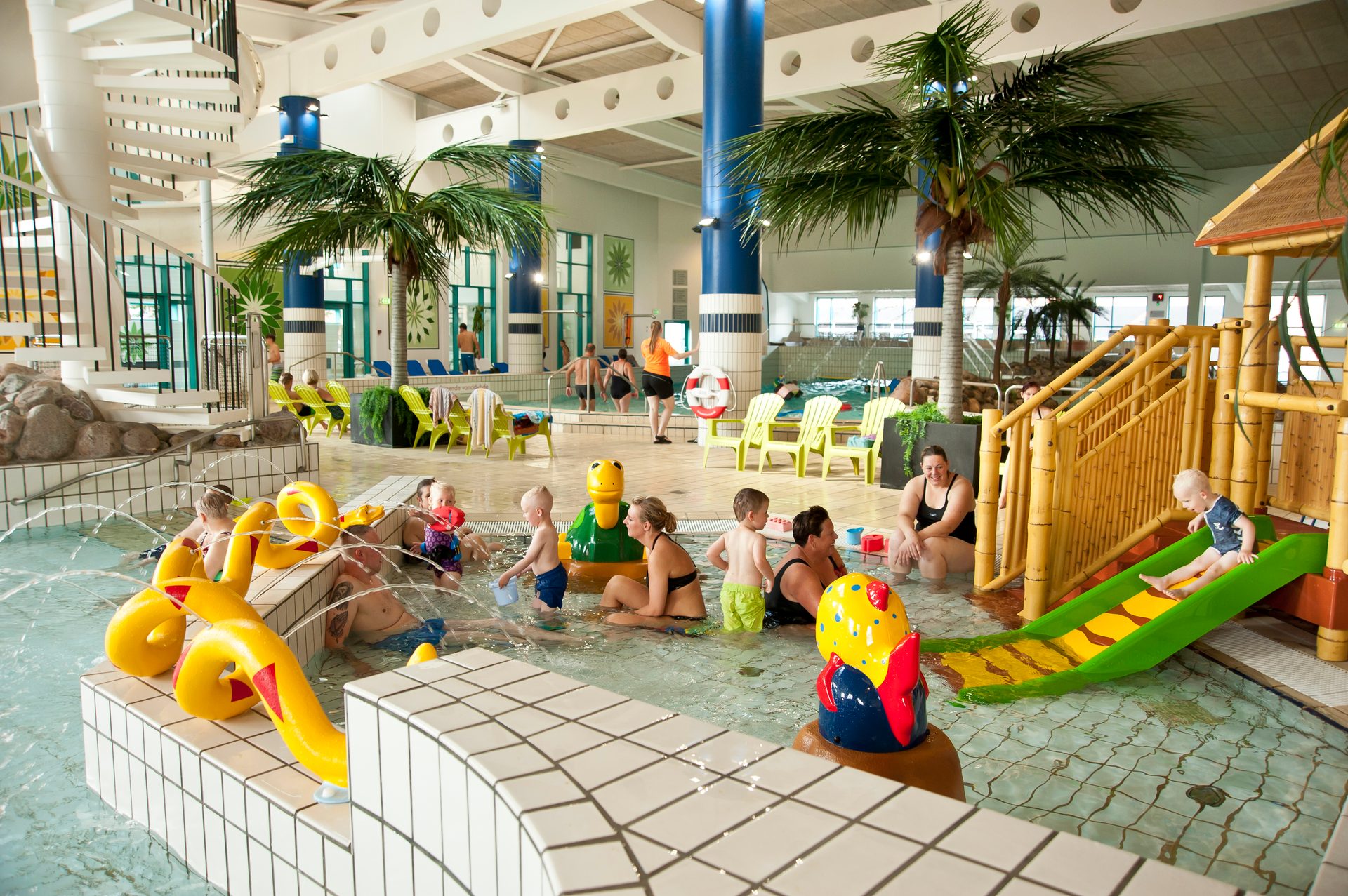A busy indoor water park with families playing in shallow pools, featuring slides, water features, and palm trees.