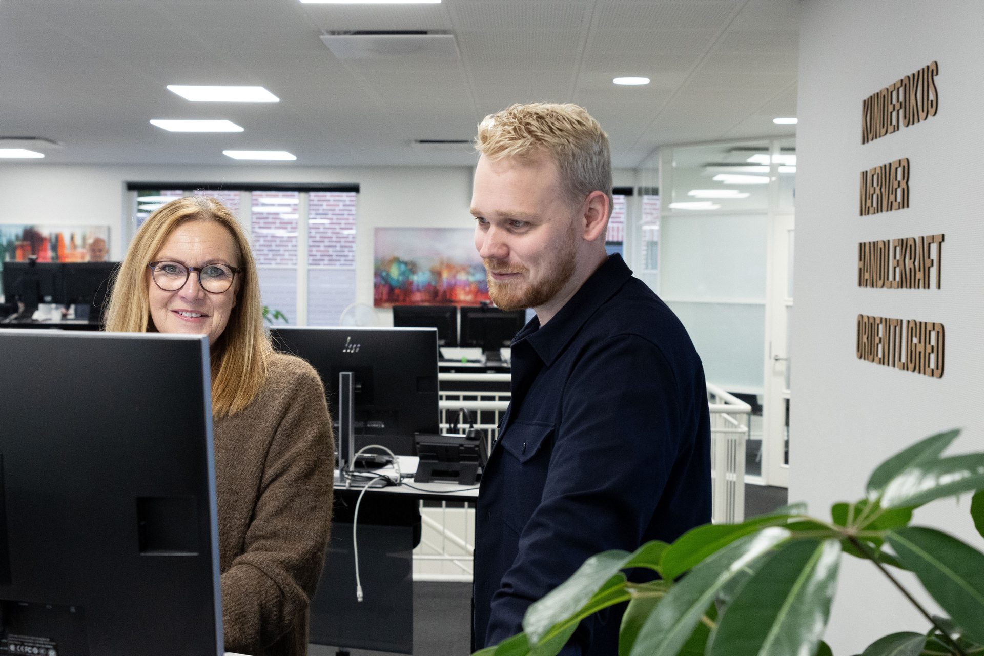 Two colleagues look at a computer in an office, a woman with glasses smiles.