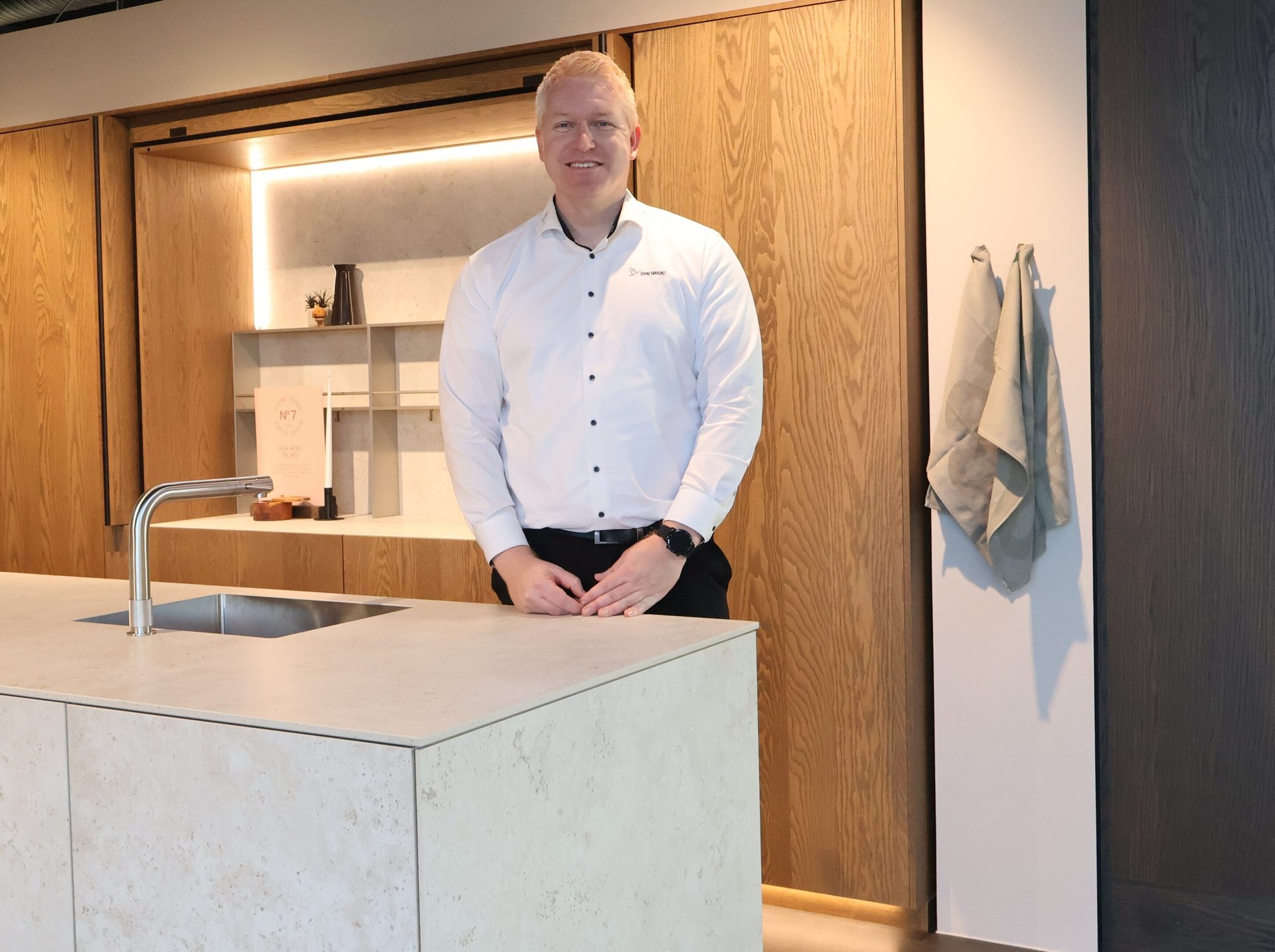 A smiling man in a white shirt stands behind a light-colored kitchen island with a sink and faucet.
