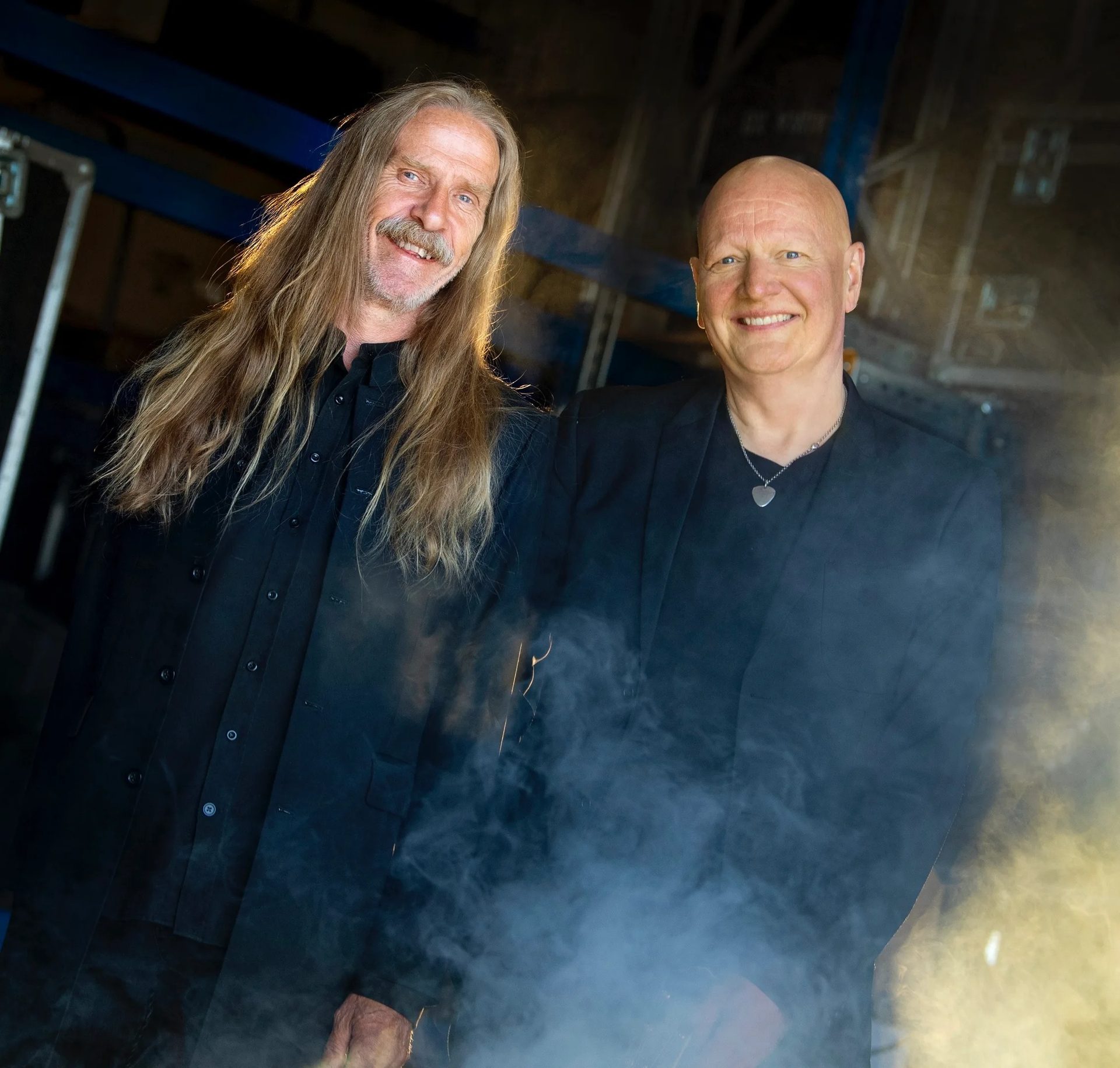 Two smiling men, one with long hair and beard, the other bald, pose in dark clothing amidst stage smoke.