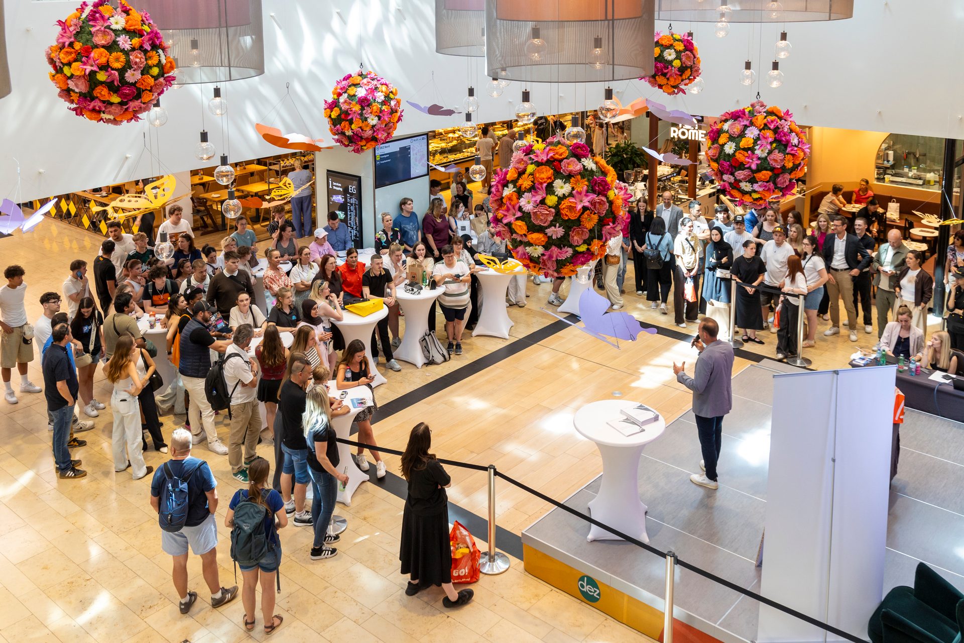 Indoor event with a speaker on stage, large crowd, and colorful floral decorations.