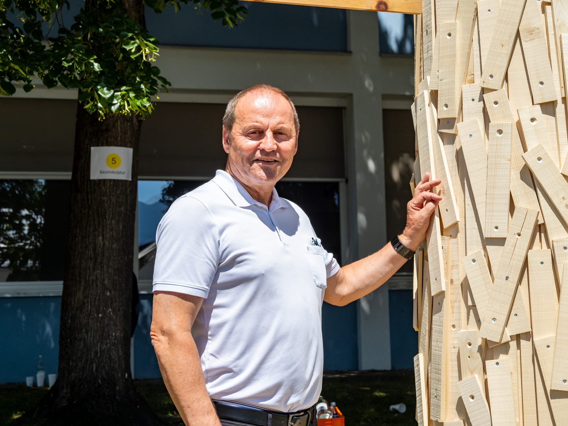 A man in a white polo shirt stands smiling next to a textured wooden sculpture outdoors.