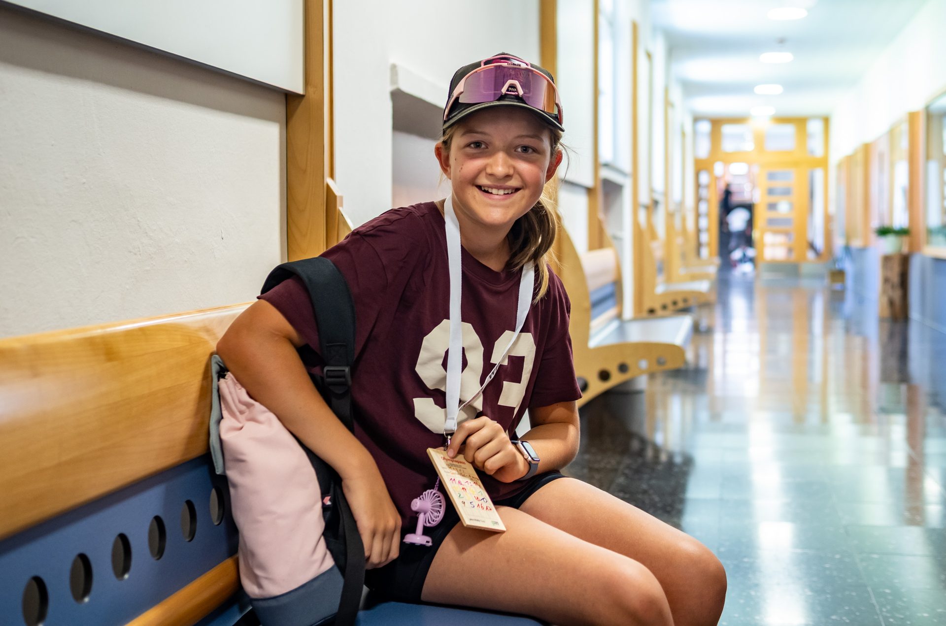 Smiling girl wearing a cap and t-shirt, sitting in a school hallway with a backpack.