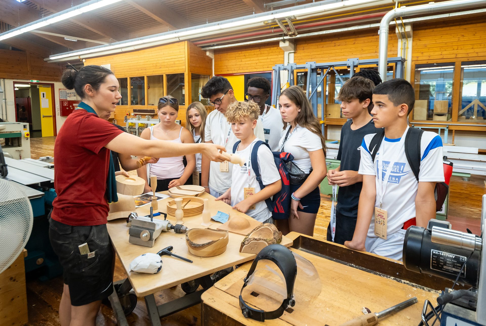 Instructor shows wooden crafts to diverse teenagers in a woodworking workshop.