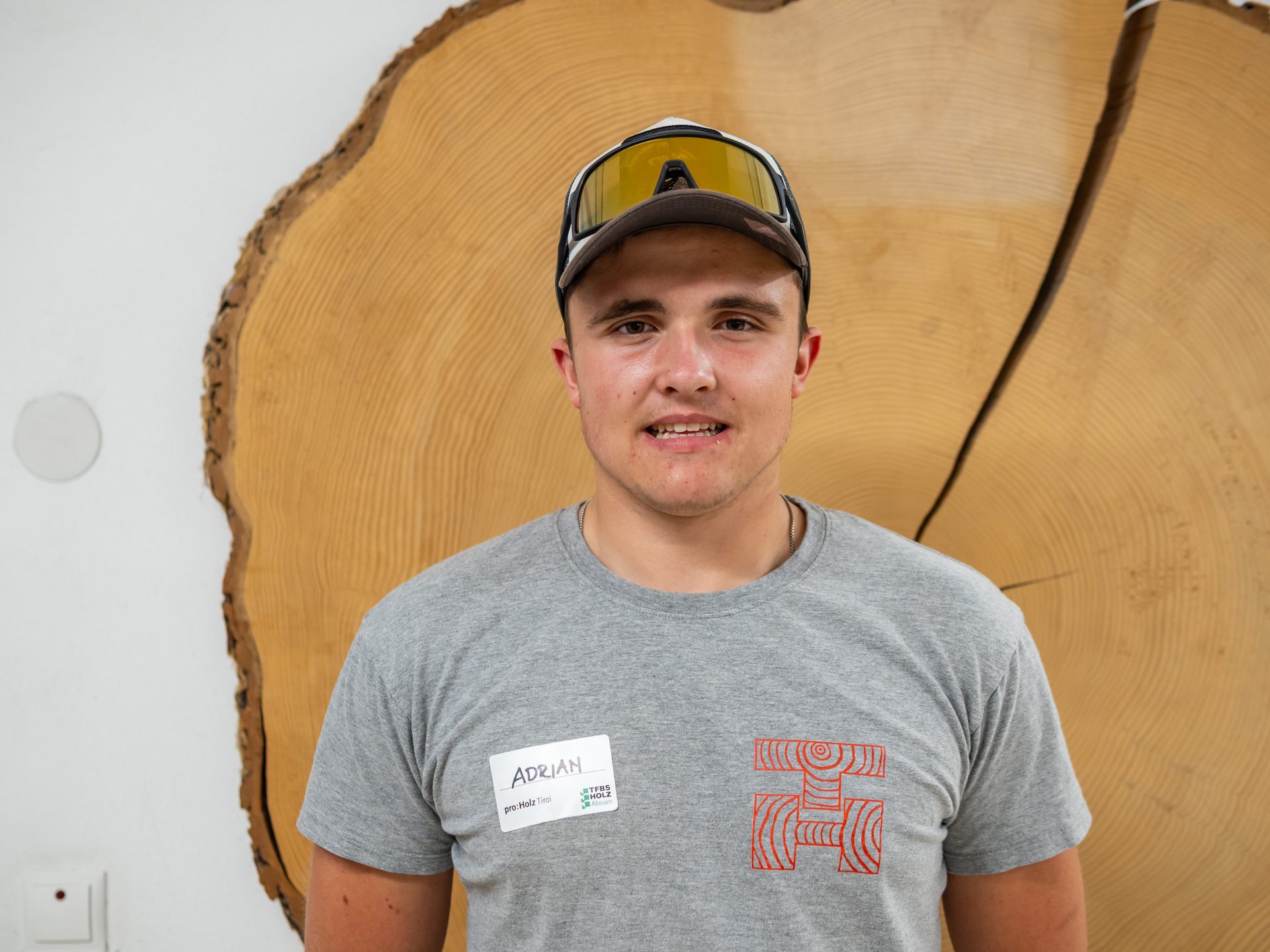 Young man Adrian with a nametag smiles, wearing a grey t-shirt and cap, in front of a wooden tree ring slab.