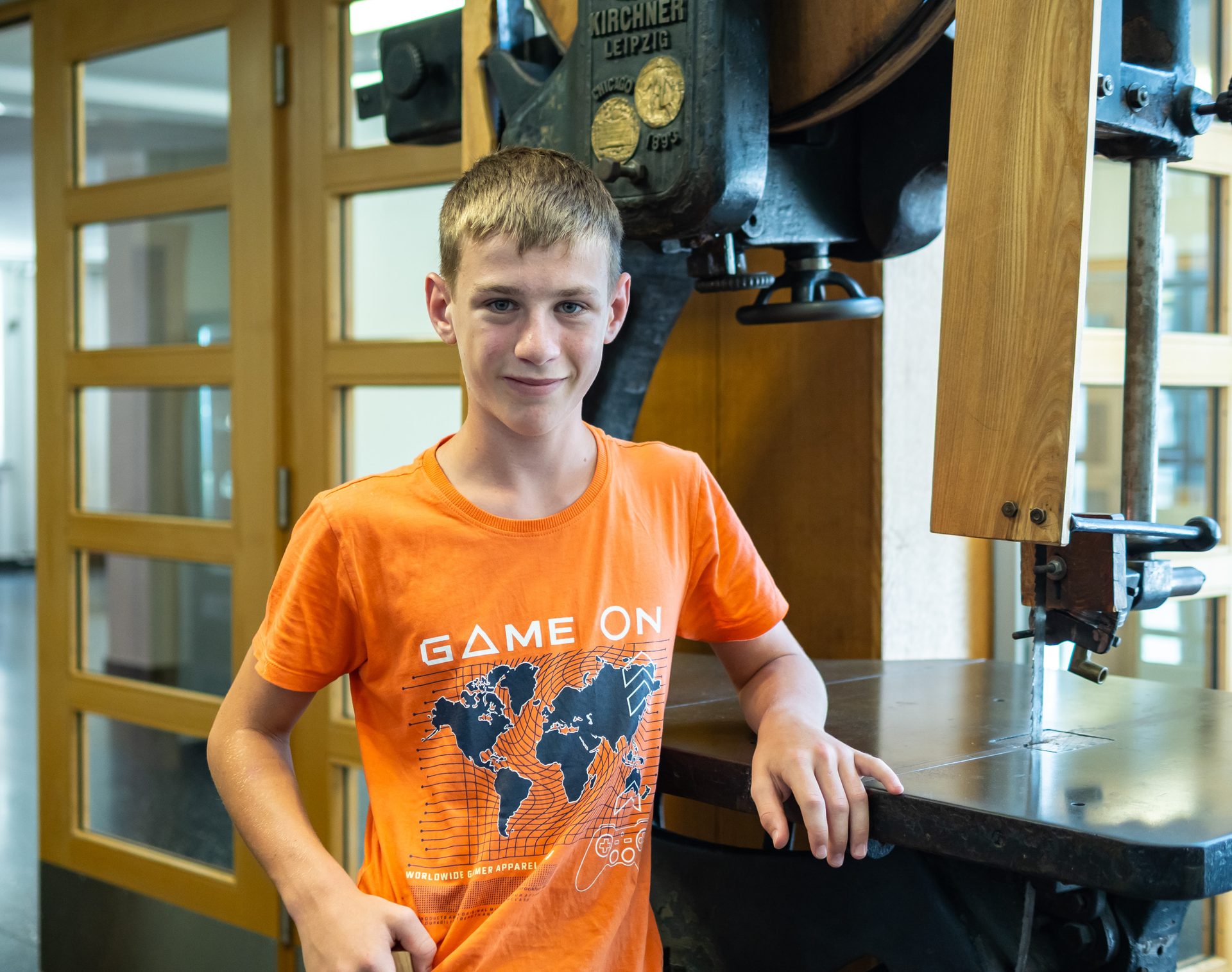 A young boy in an orange "GAME ON" t-shirt smiles while standing next to an old band saw machine.