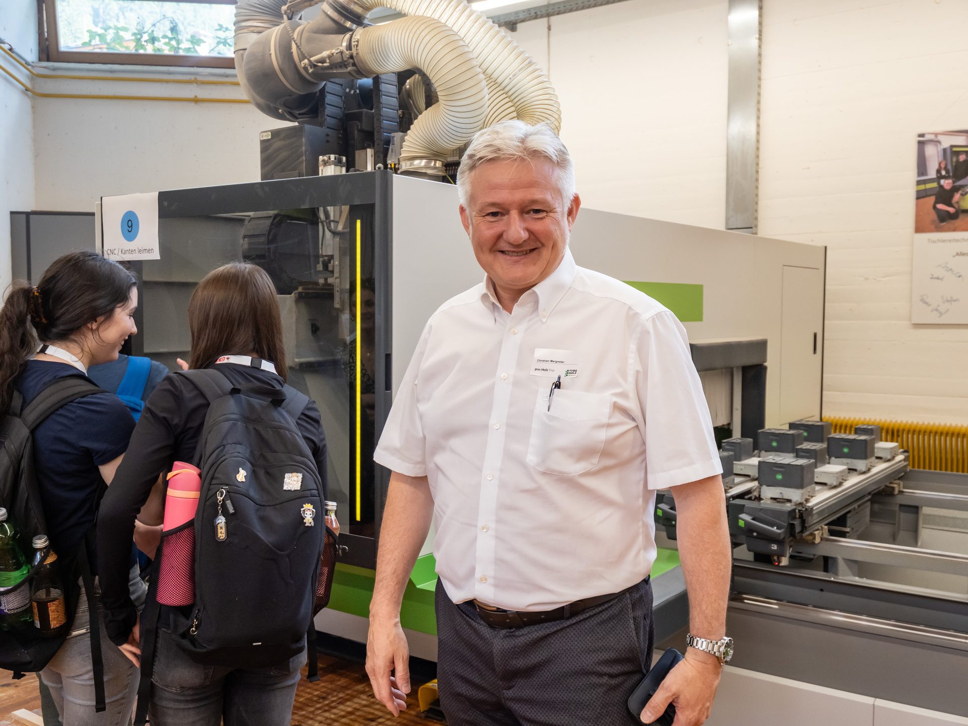Smiling man in a white shirt with a name tag, standing in a workshop with two women and a large machine.