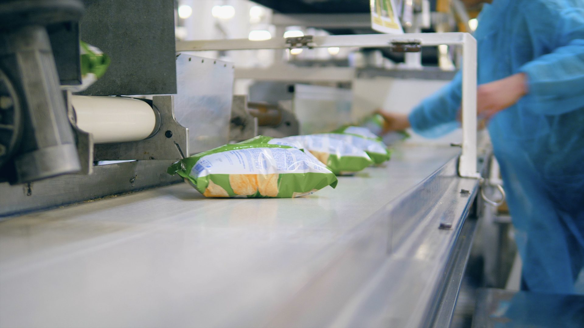 A person packs bags with potato crisps into a box, taking them from a conveyor