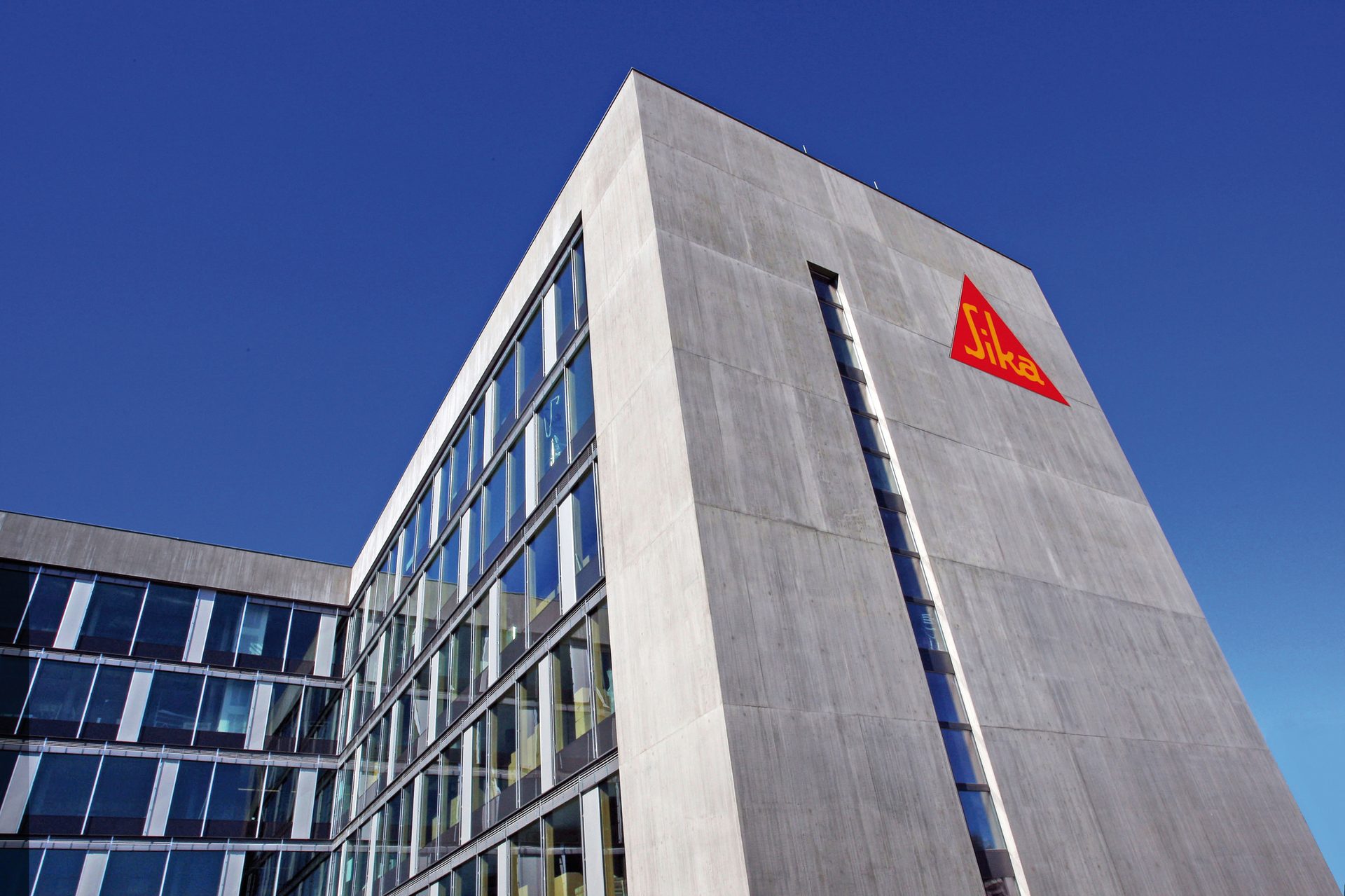 Modern concrete building with large glass windows and the red Sika logo under a clear blue sky.