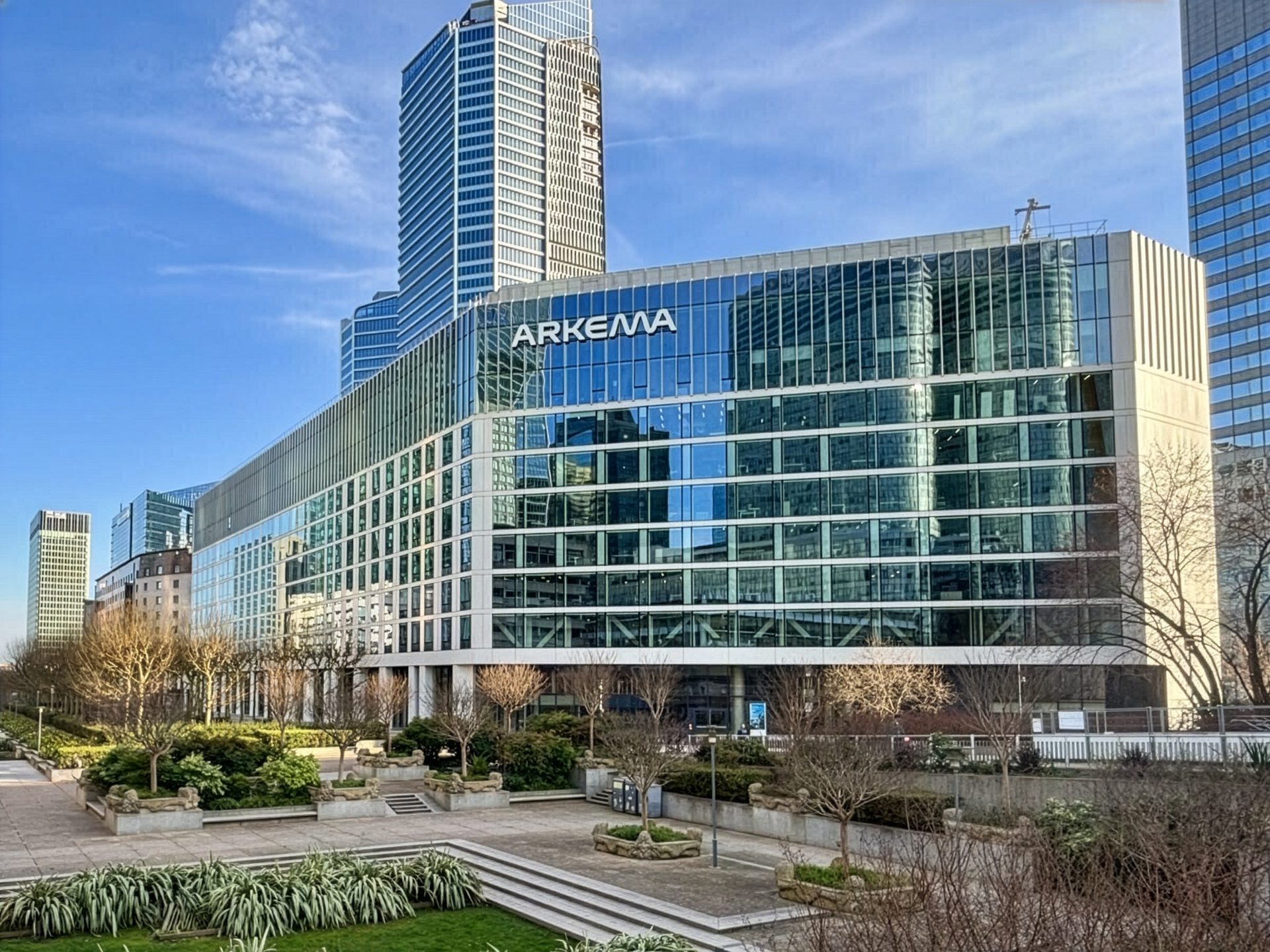 Modern glass Arkema office building with a green park in the foreground under a blue sky.