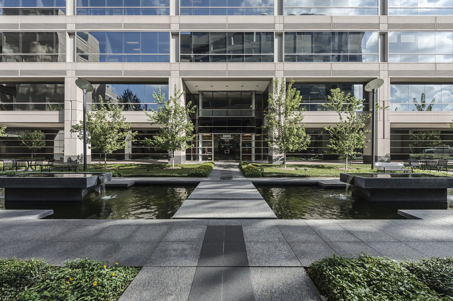 Modern building entrance with reflective windows, flanked by green trees and a symmetrical water garden with pathways.