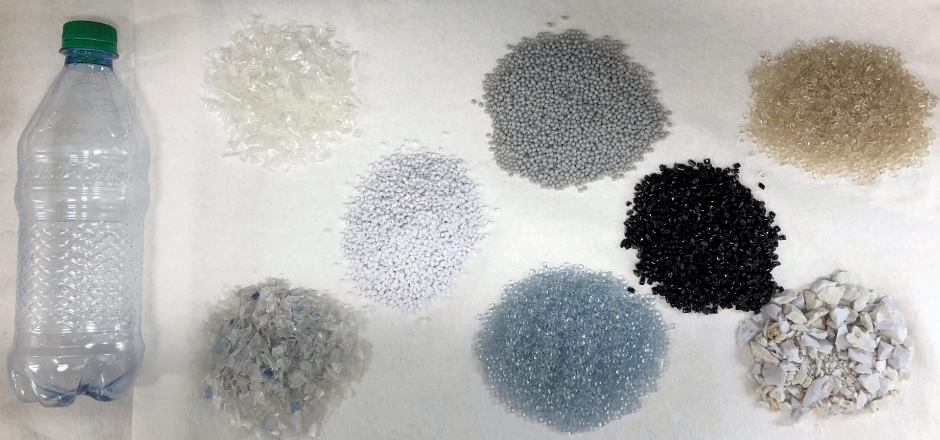 A plastic bottle next to piles of various plastic materials (flakes, pellets, chunks) in different colors.