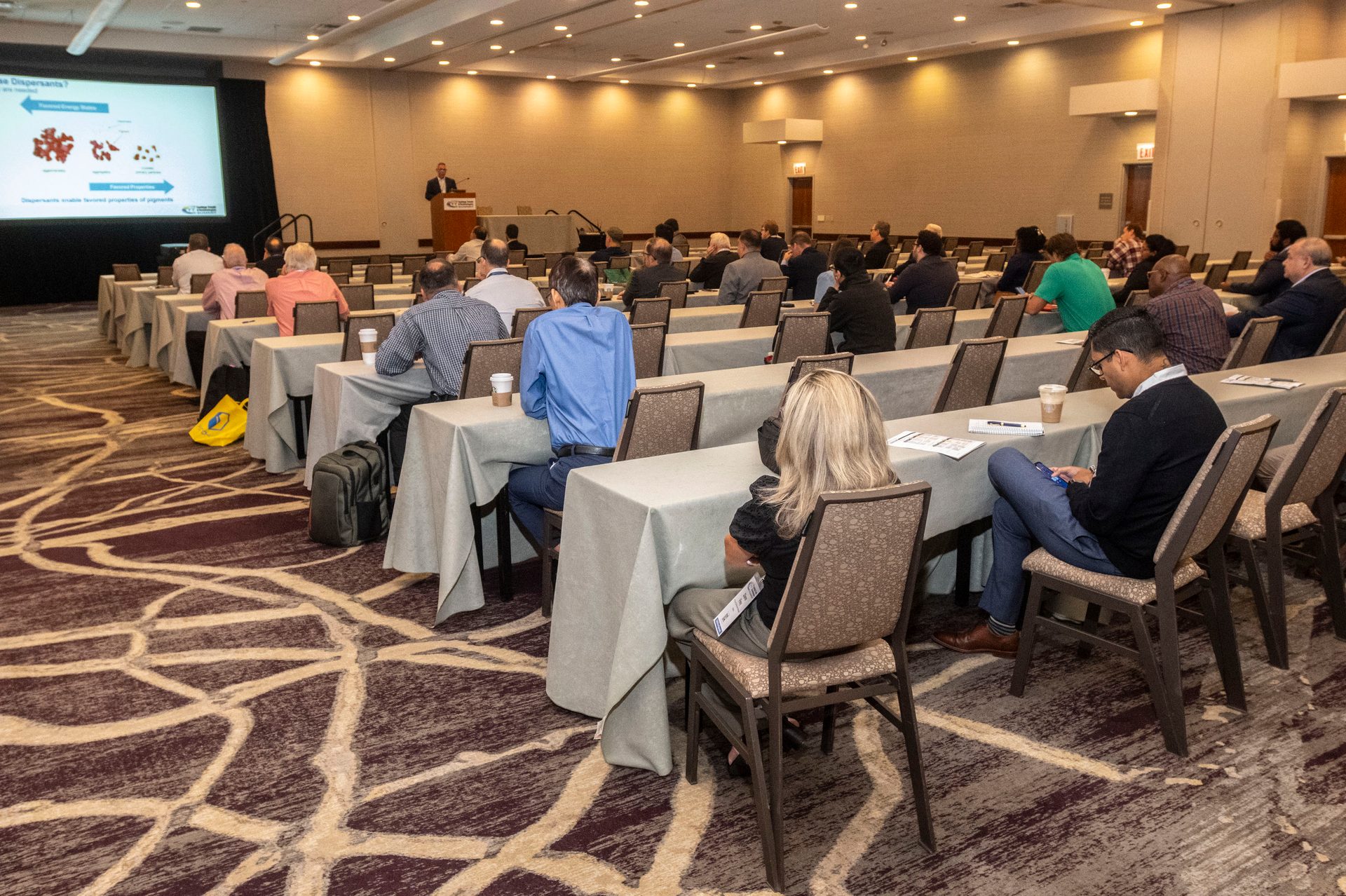 A speaker presents to an audience seated at tables in a conference room, with a large screen displaying content.