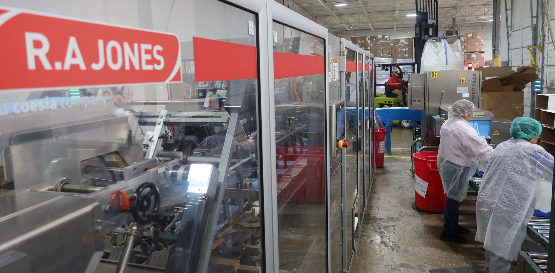 Image of workers using the new handload cartoner solution from R.A Jones that features a configurable bucket conveyor length, allowing customers to tailor the machine's footprint and speed to their specific needs.