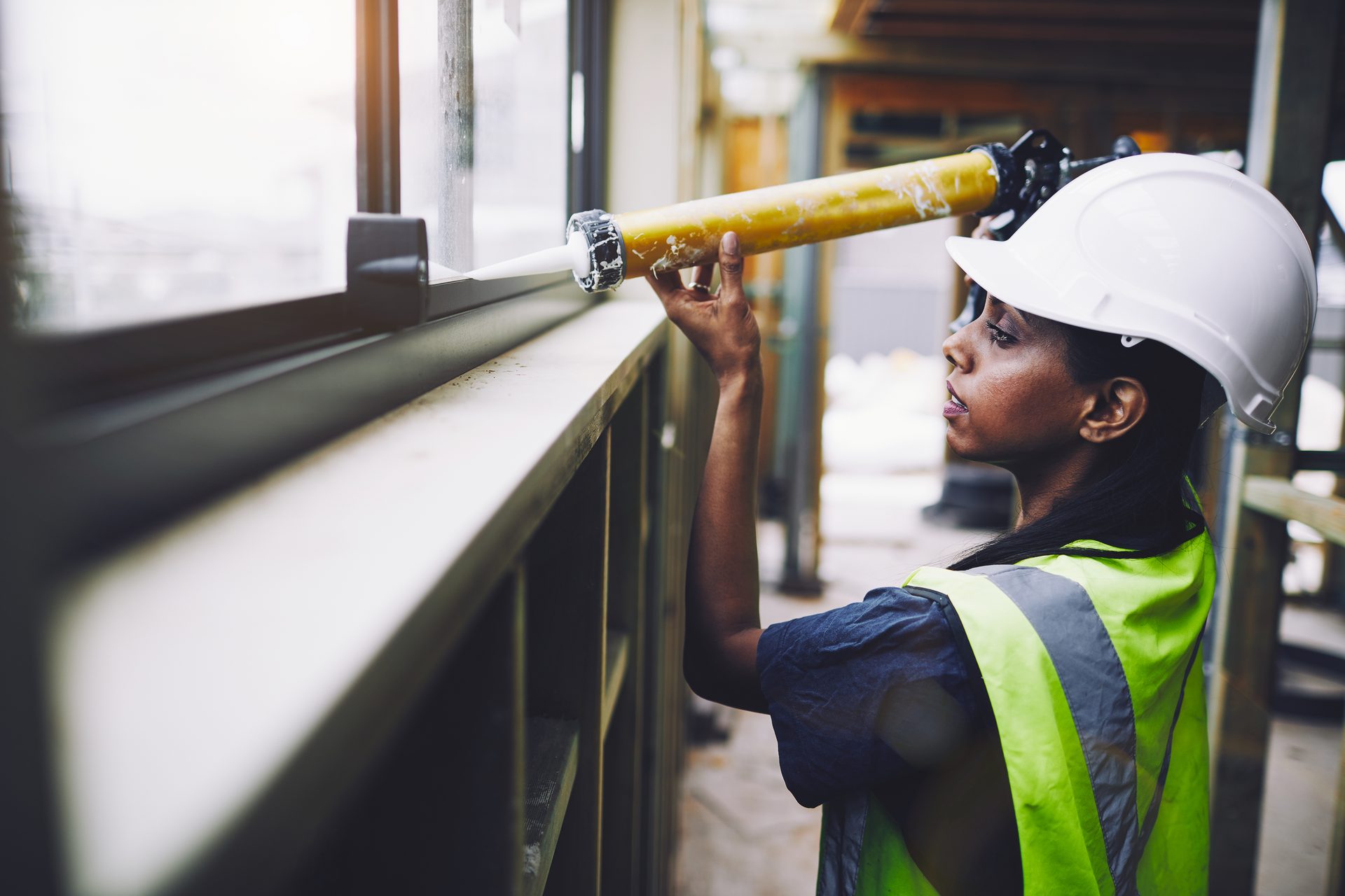 Woman in hard hat and hi-vis vest caulking a window.