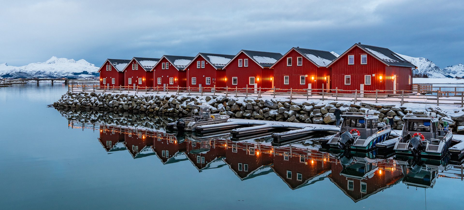 Red rorbuer cabins line a snowy harbor reflecting in still water with mountains