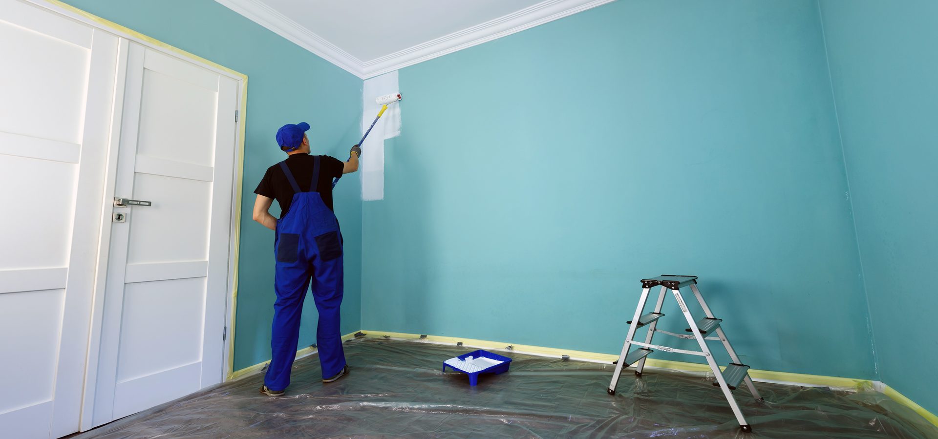 Painter in blue overalls painting a light blue wall white with a roller.
