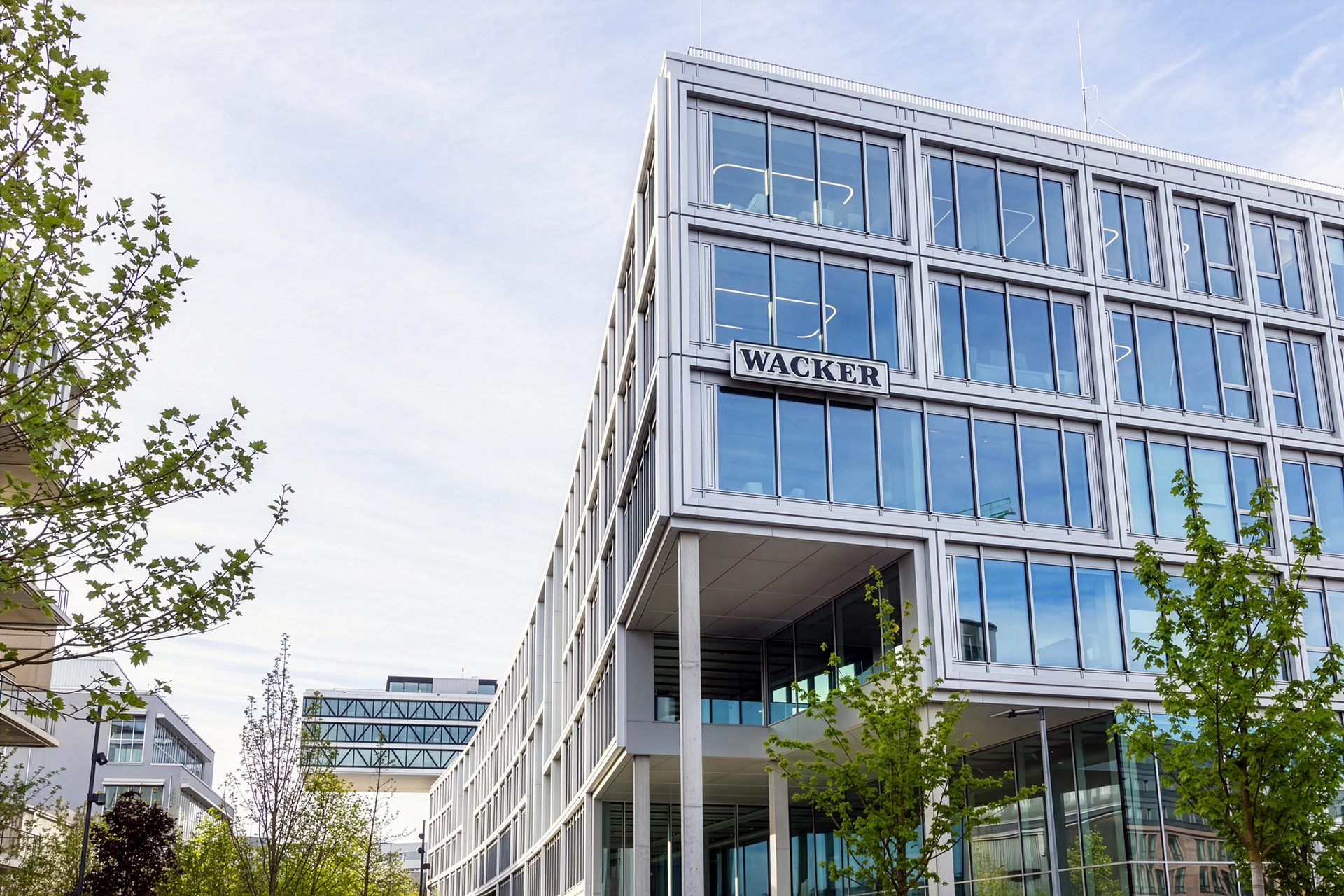 Modern office building with large glass windows and a prominent "WACKER" sign, flanked by green trees.