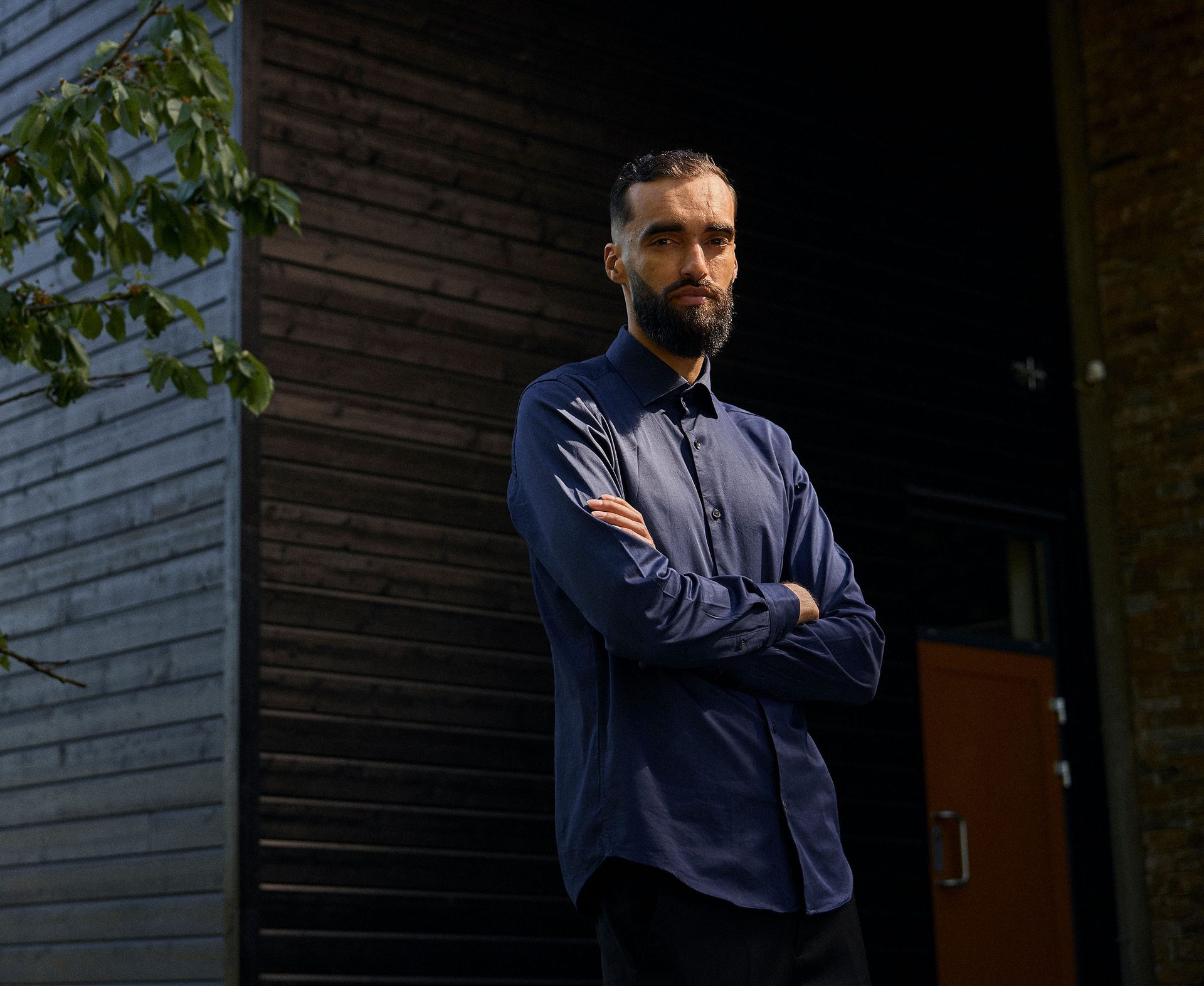 Man with beard in dark shirt, arms crossed, standing outside a dark wooden building.