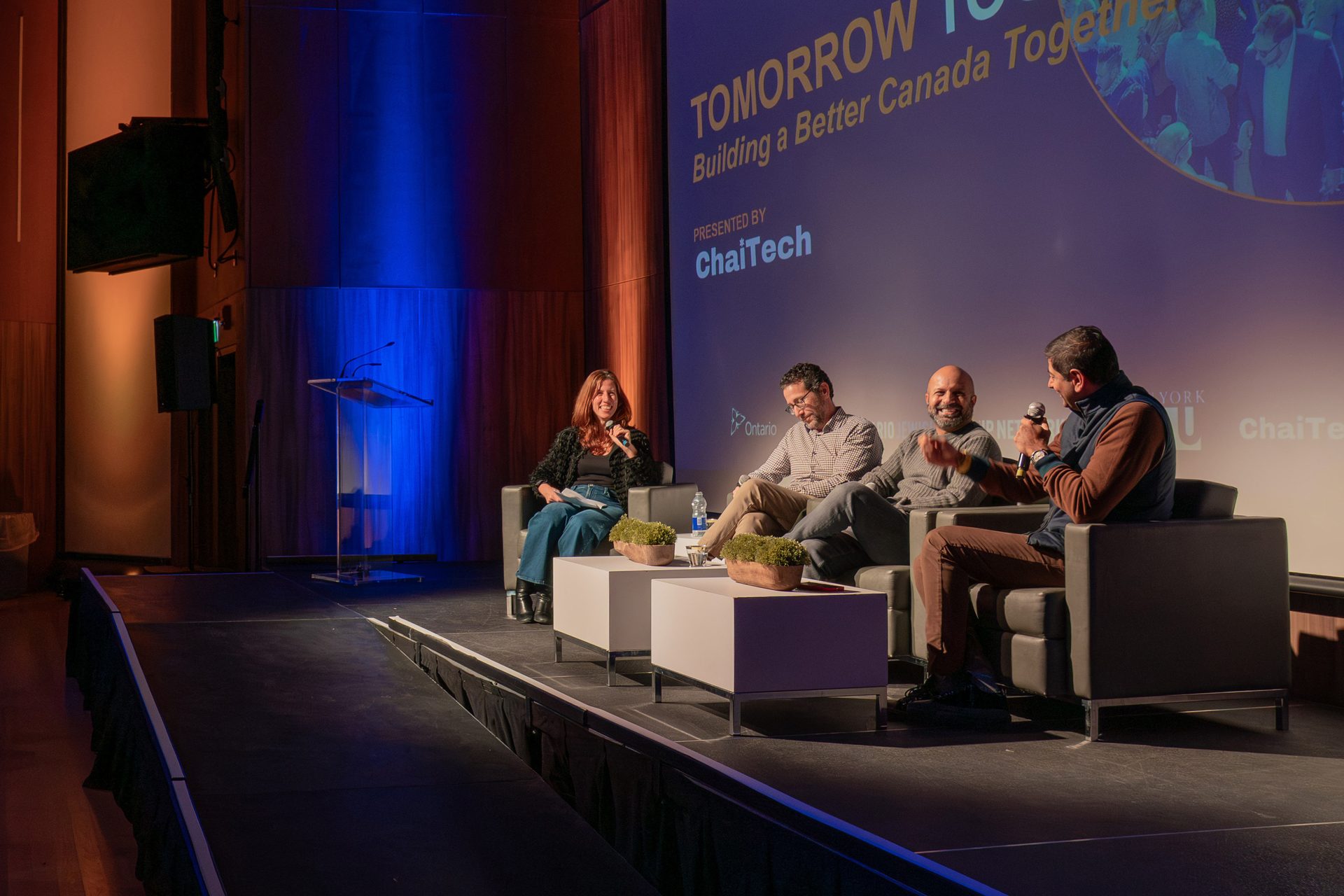 A panel discussion on a stage with four speakers, a large screen, an audience, and colorful lighting.