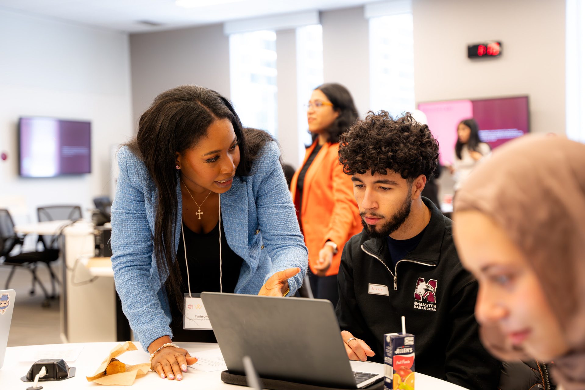Woman in blue blazer helps man with laptop; others work in a bright, modern room.