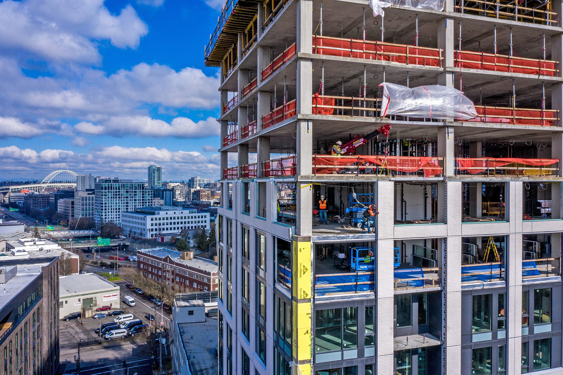 A tall building under construction with workers and equipment, against a city skyline with a bridge.