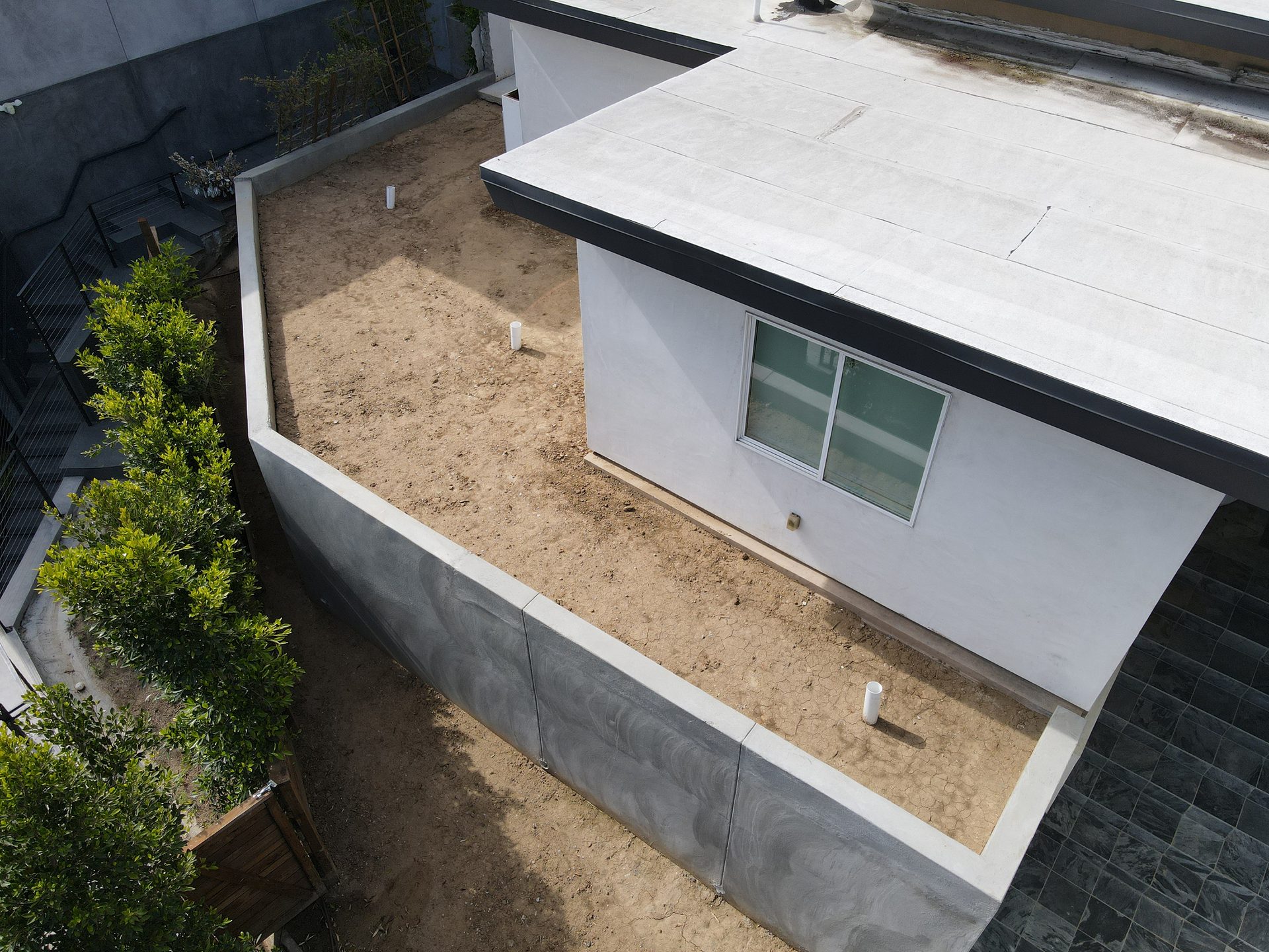 Aerial view of a barren backyard with a white building, concrete walls, green hedges, and dirt. 