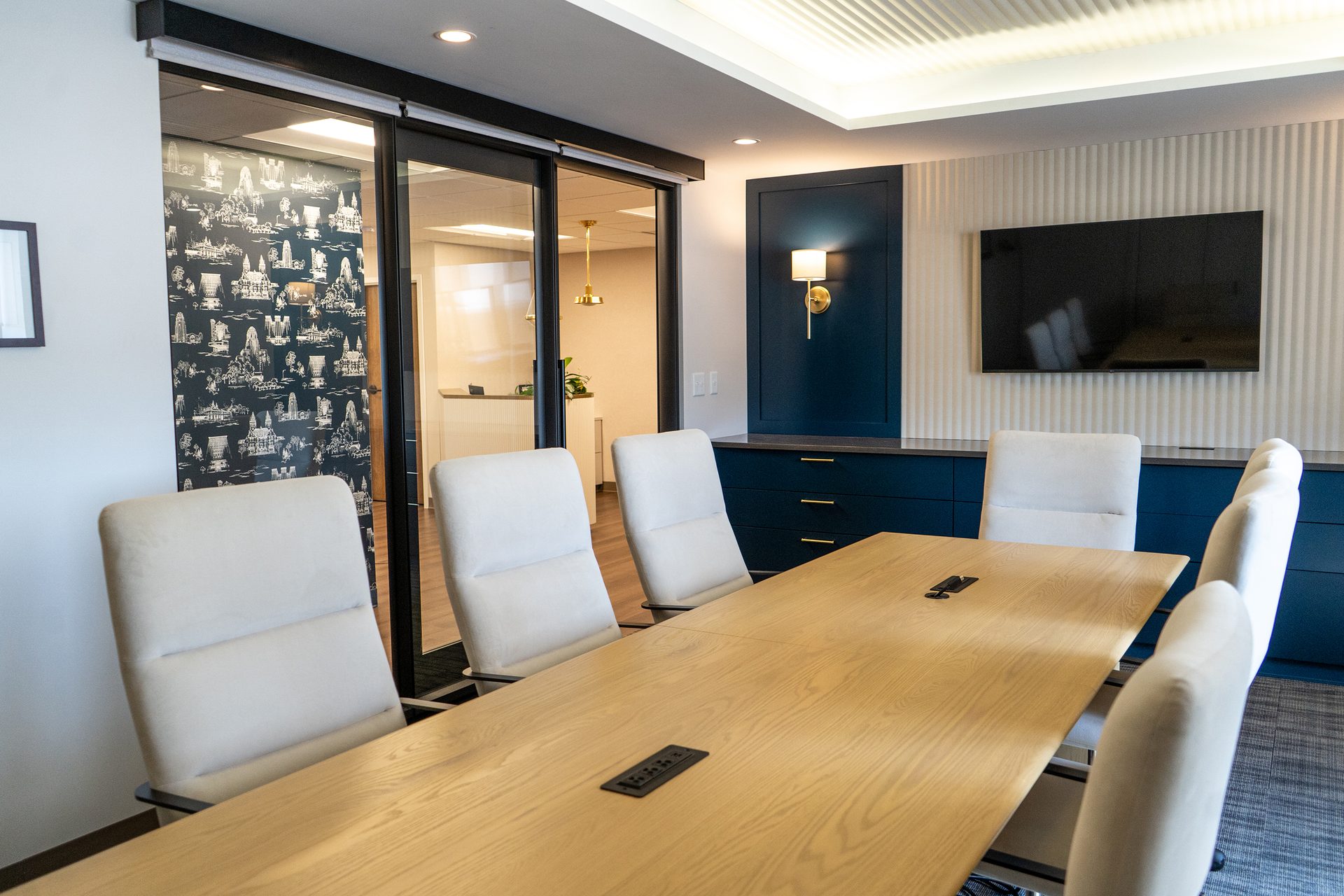 Conference room with long wooden table, cream chairs, blue cabinet, TV, and glass doors.