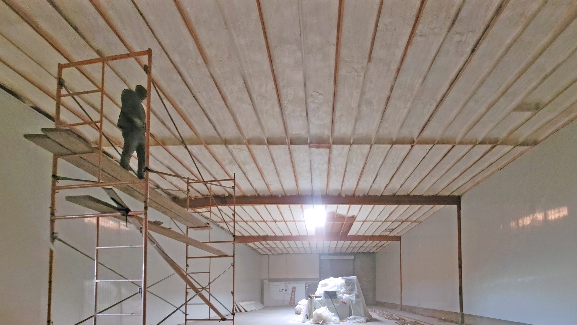 Worker on scaffolding in a large room under construction, featuring an exposed ceiling and bright skylight.