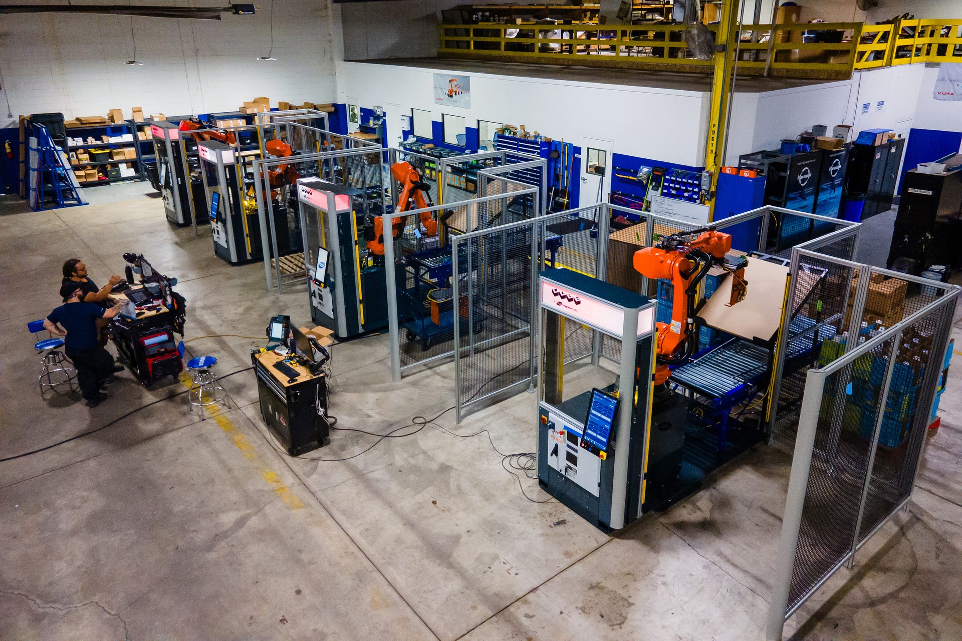 Overhead view of an automated factory floor with multiple robotic work cells and two technicians.