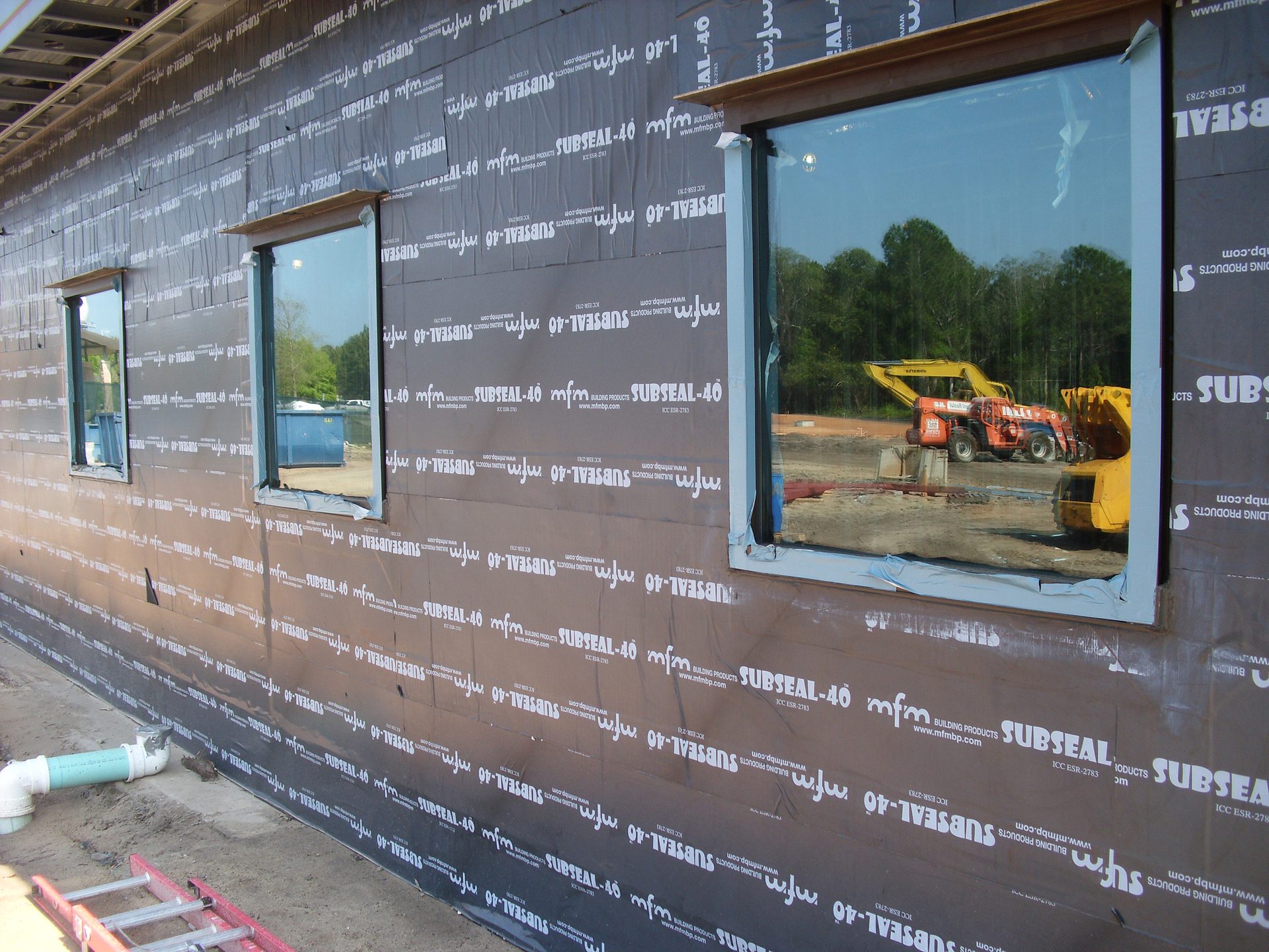 Building wall covered in black "SUBSEAL-40" membrane with windows reflecting construction equipment.