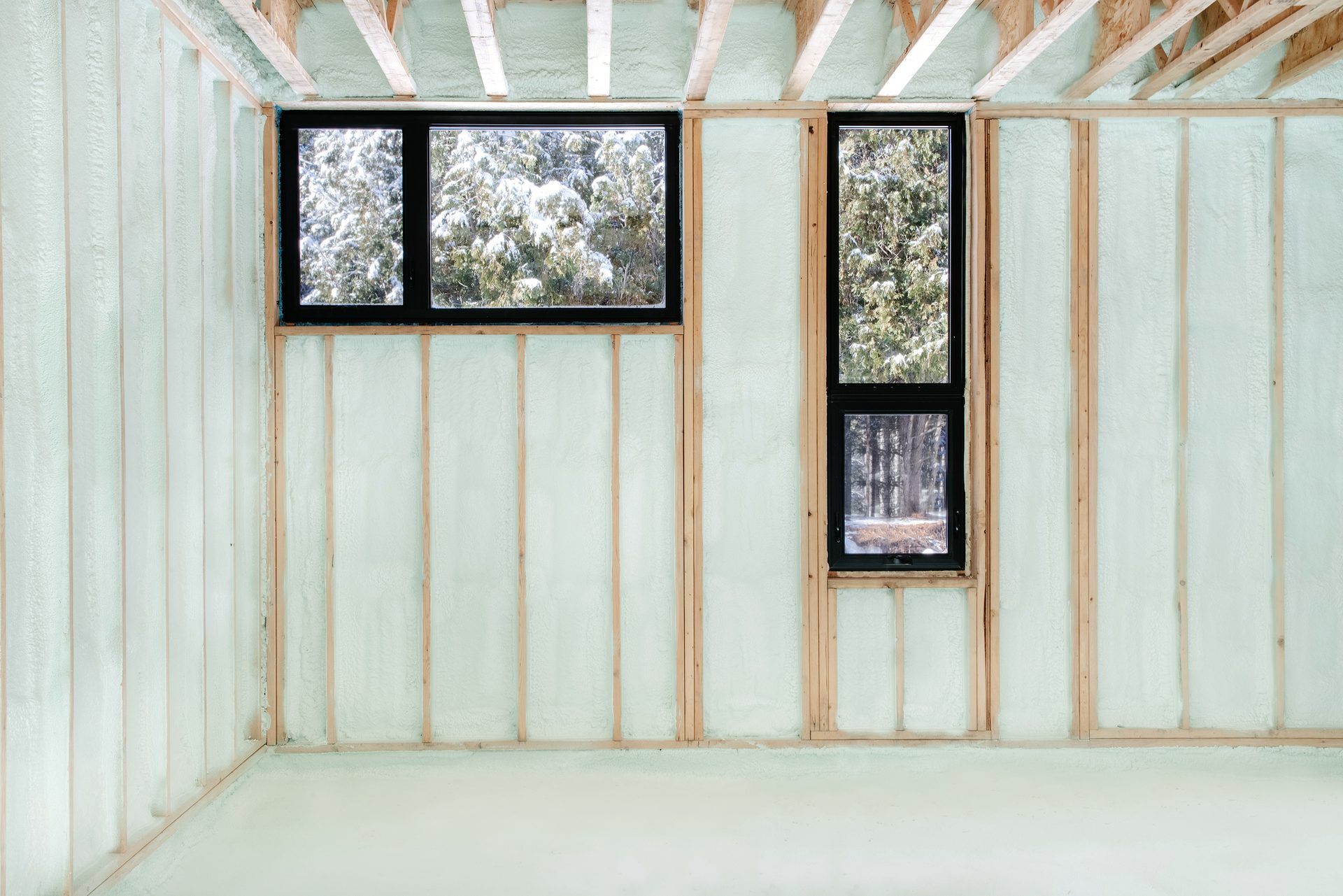 Interior of a room under construction with light green spray foam insulation, wood framing, and snowy view.
