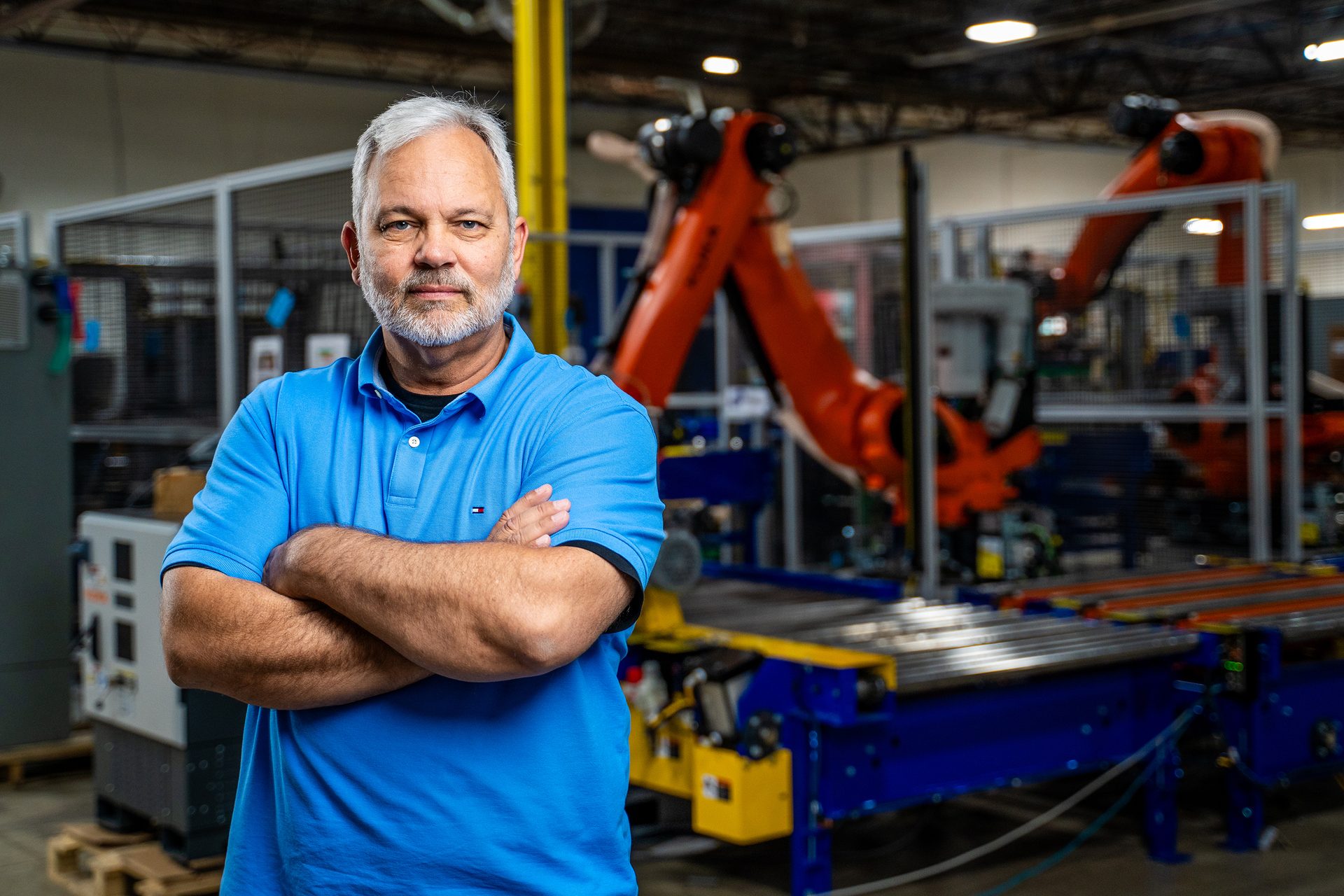 Gray-haired man in blue polo shirt, arms crossed, in an industrial facility with robots.