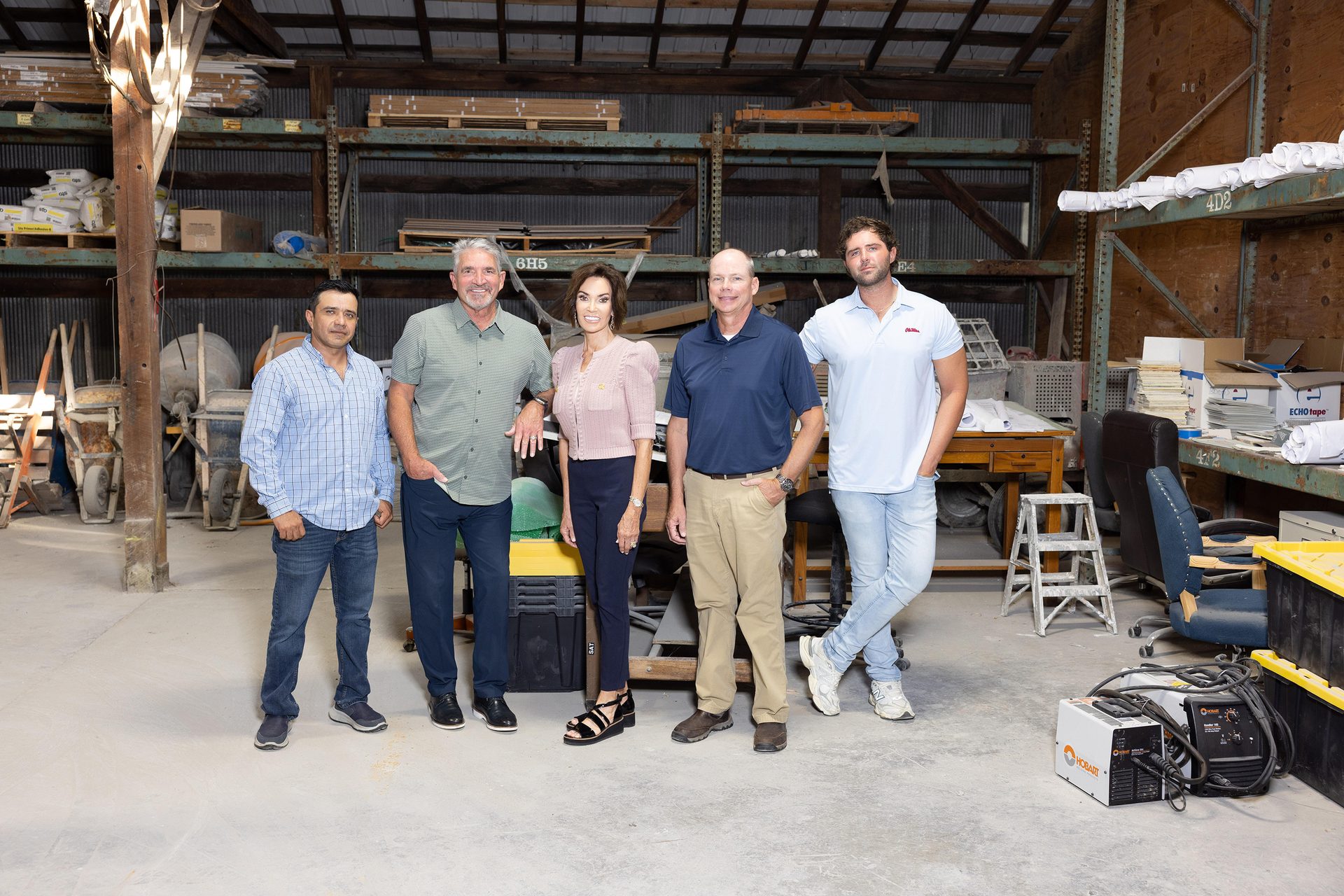 Five smiling people (four men, one woman) pose in a warehouse with shelving.