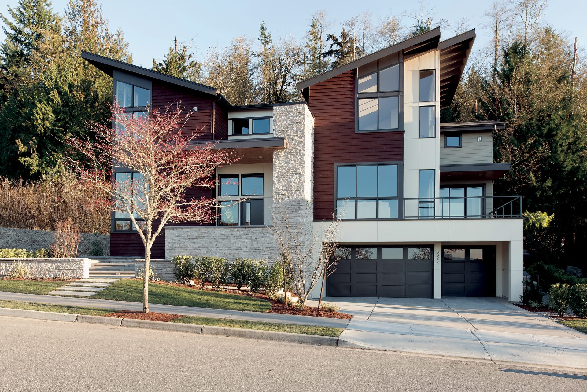 Modern house with red siding, stone facade, large windows, and a multi-level design amidst trees.