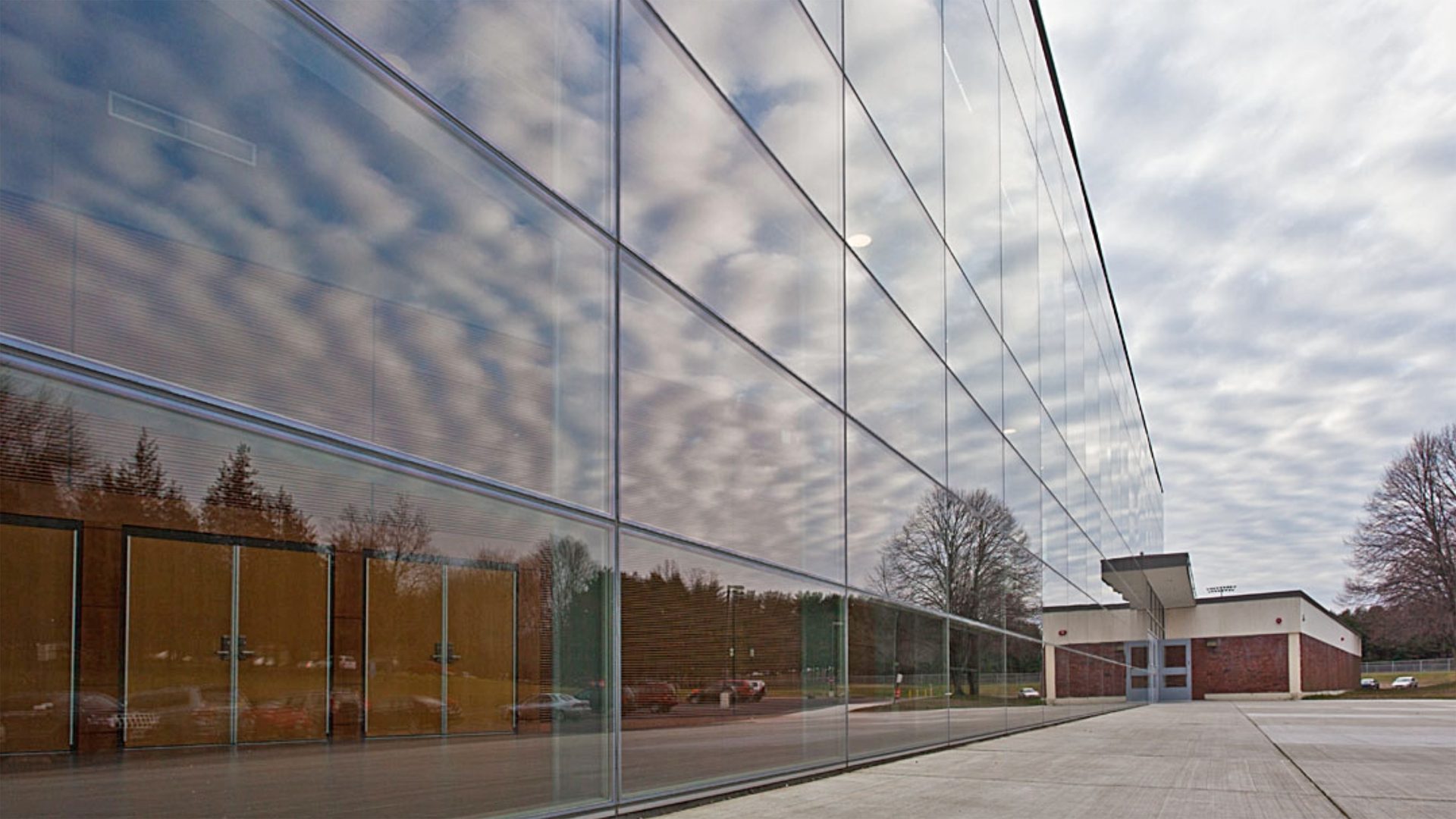 Glass building reflecting cloudy sky, trees, and an adjacent brick structure.