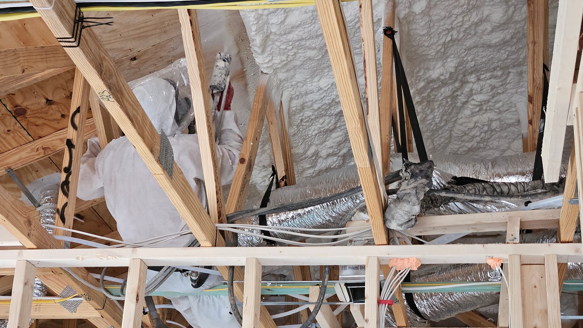 Worker in protective suit applying spray foam insulation to an attic roof structure.