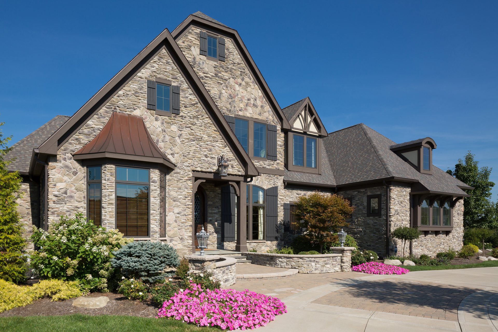 Grand stone house with multiple gables, a copper-roofed bay window, and vibrant pink flowers in the foreground.