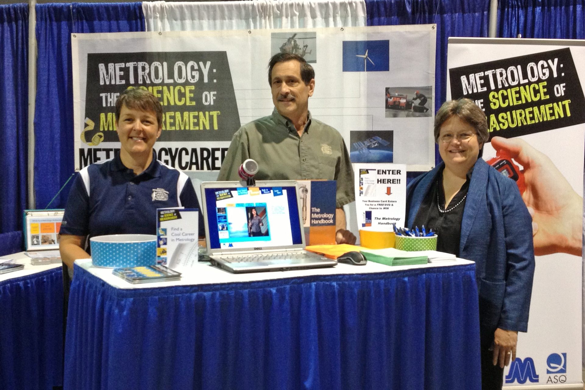 Three individuals staff a Metrology booth with educational banners and promotional materials.